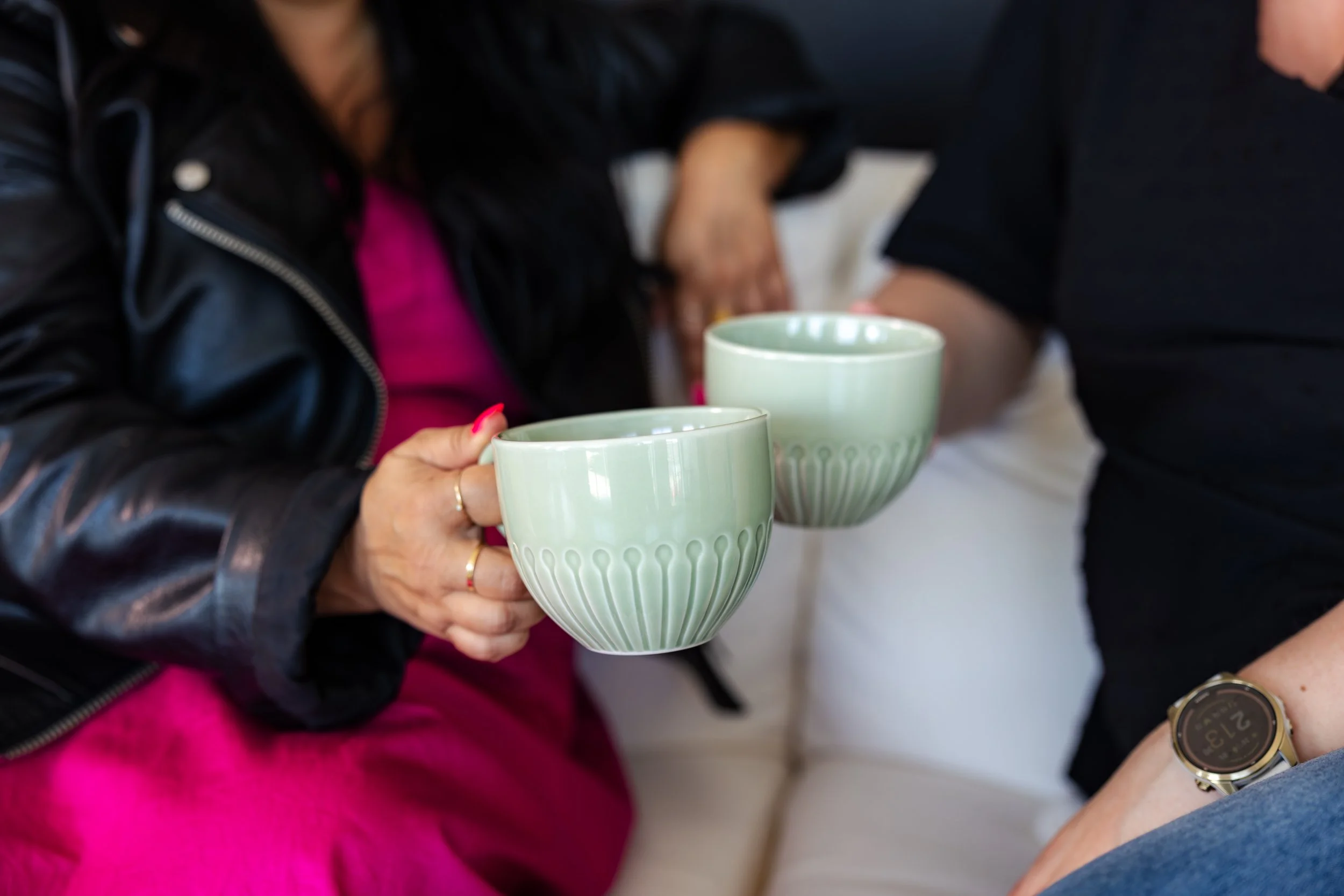 Deux personnes tiennent des tasses vert pastel lors d'une rencontre, avec des vêtements colorés et une montre visible.