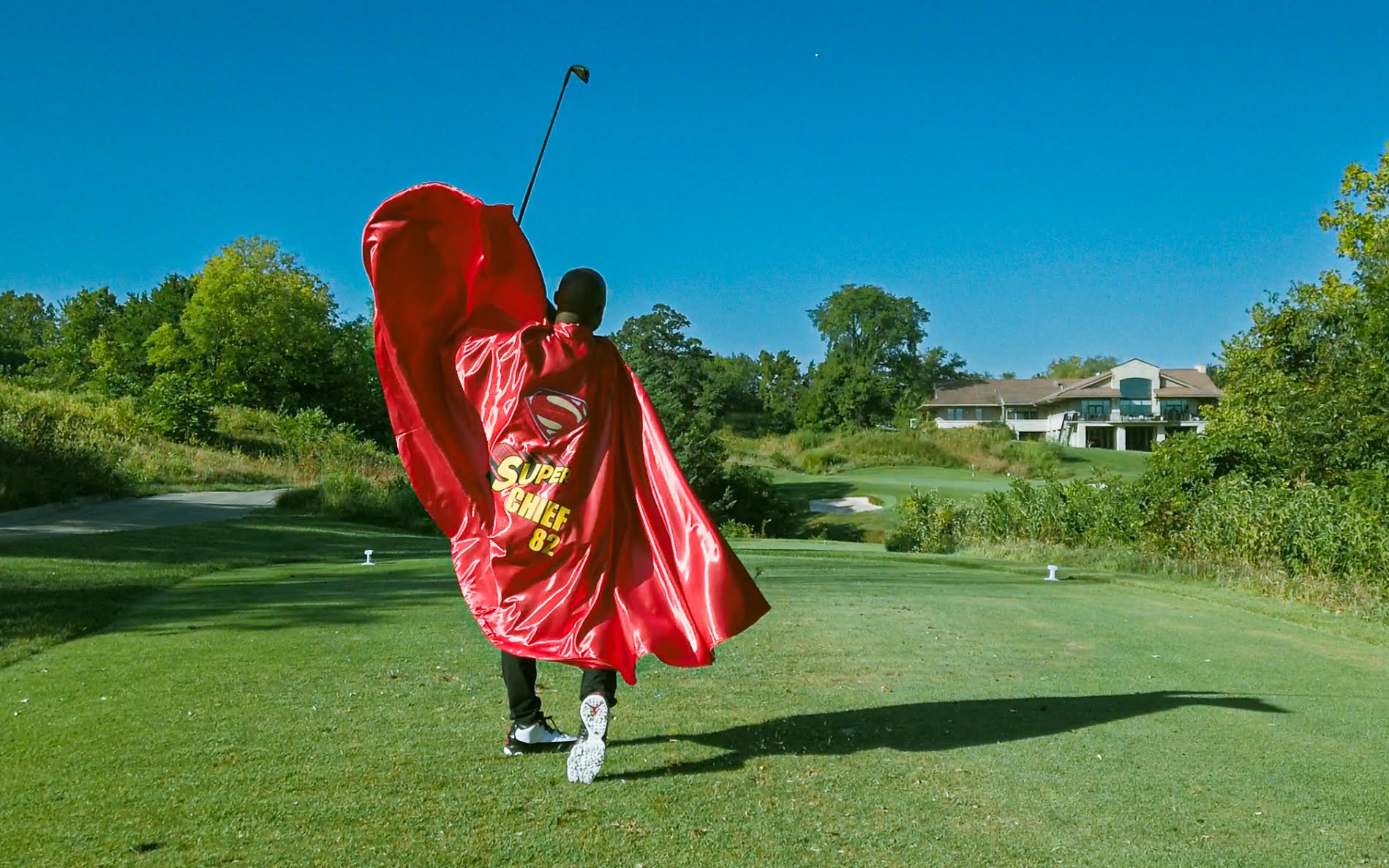 A person dressed as Super Chief, wearing a red cape with a Superman emblem and the words 'Super Chief 82,' swings a golf club on a golf course under a clear blue sky, with trees and a house in the background.