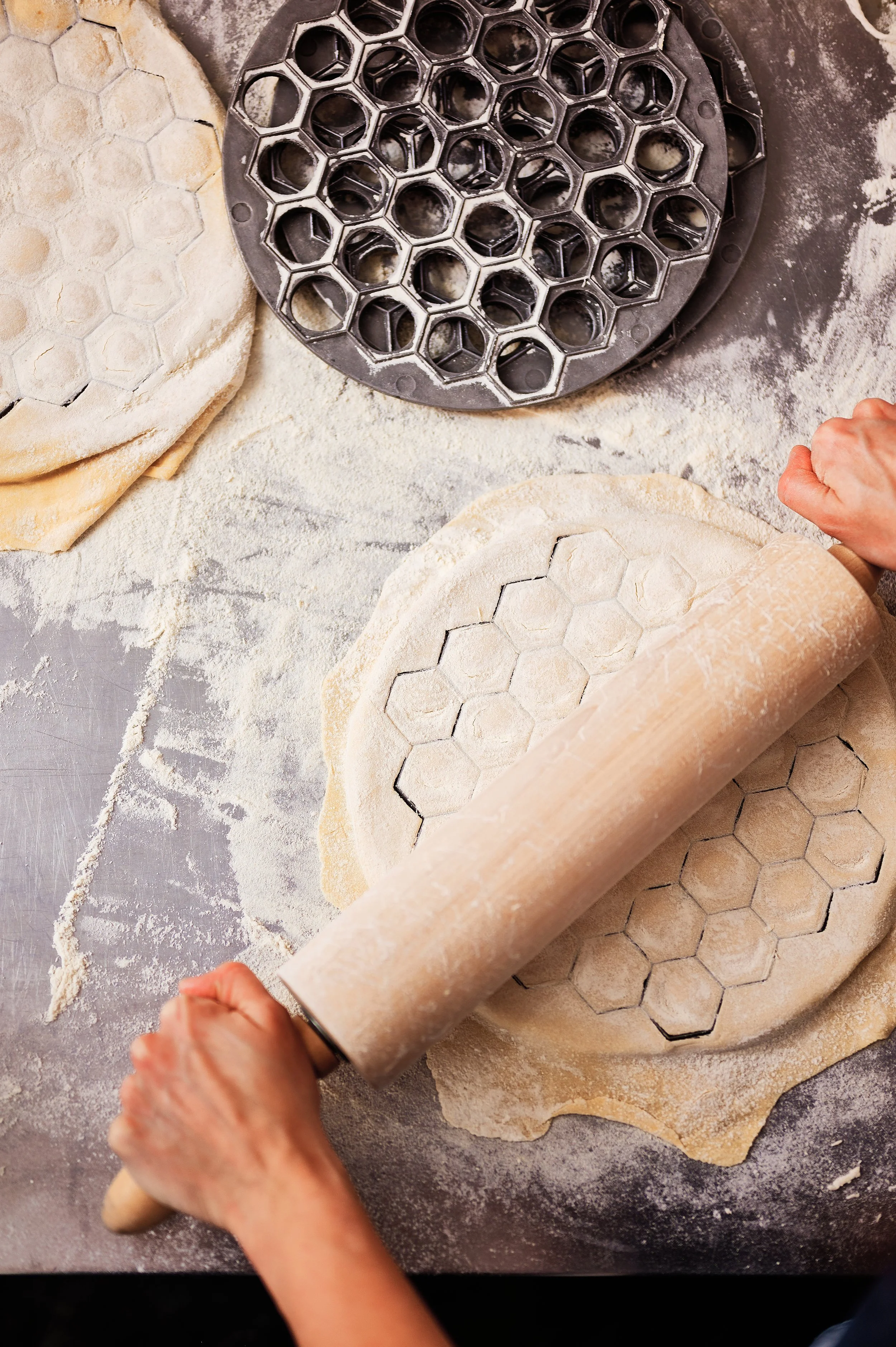 Person using a rolling pin to flatten dough with honeycomb-shaped cookie cutters on top, surrounded by flour on a work surface.