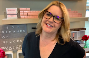 A smiling woman with blond hair and glasses, wearing a black top, standing in front of a shelf with pink boxes and a decorative sign.