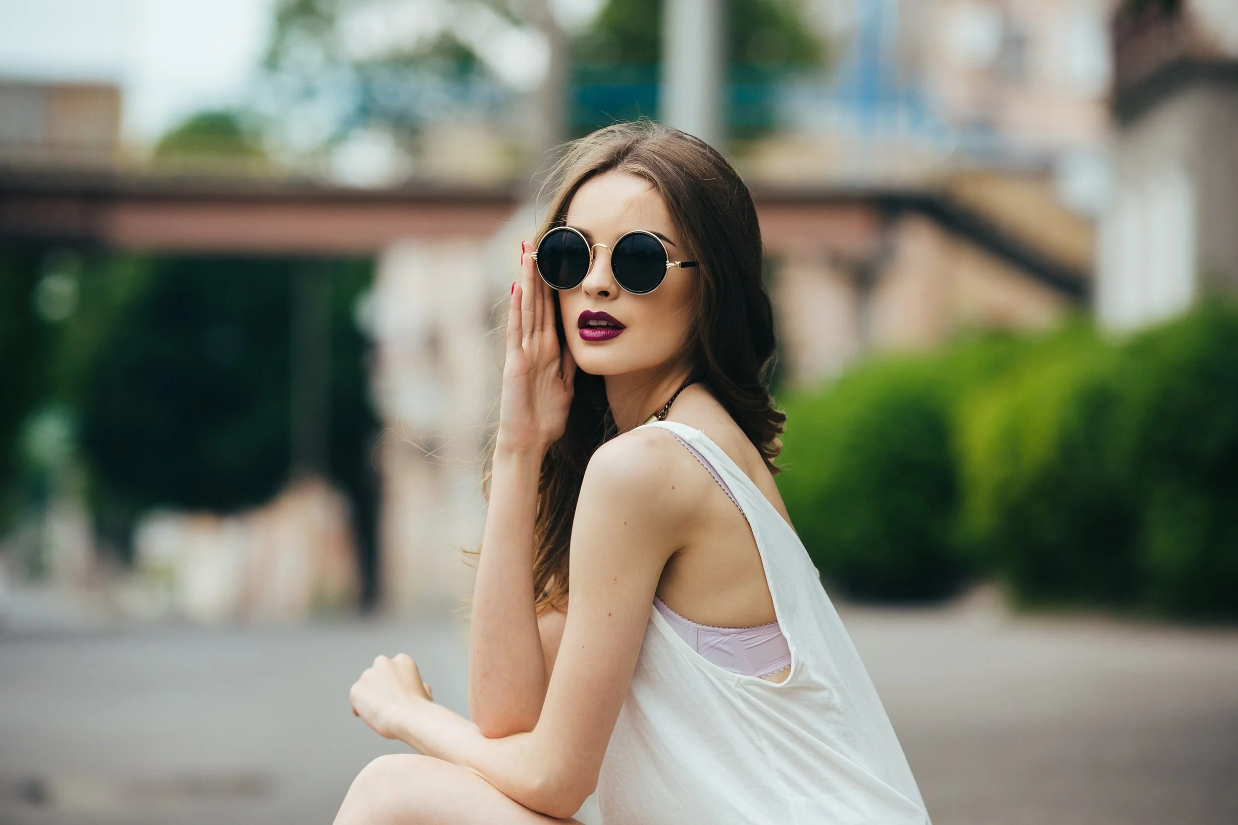 A woman with long brown hair, wearing round black sunglasses with gold frames and dark lipstick, is resting her hand on her face while looking at the camera outdoors.