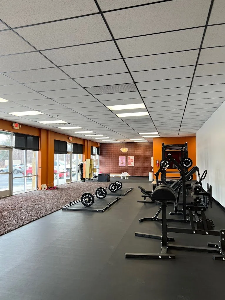 Interior of a gym in New Hope, MN with weightlifting equipment, including barbells and benches, against an orange and white wall with large windows and a carpeted area.