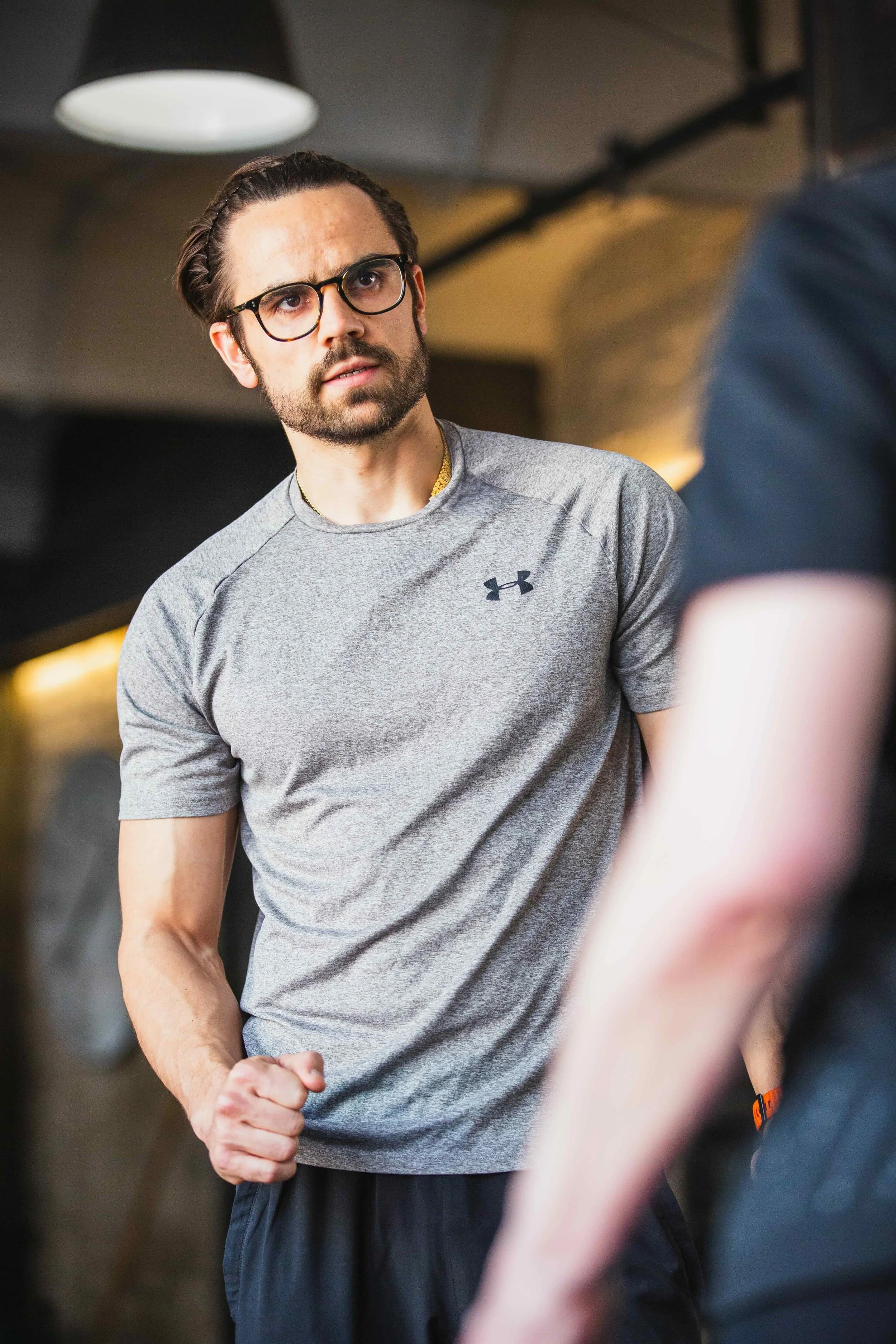 Ein Mann mit Brille und Bart, trägt ein graues Under Armour T-Shirt, sieht ernst aus und steht im Fitnessstudio.
