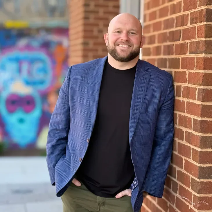 A man with a beard and shaved head smiling, wearing a blue checked blazer and black t-shirt, standing outdoors against a brick wall with colorful graffiti in the background.