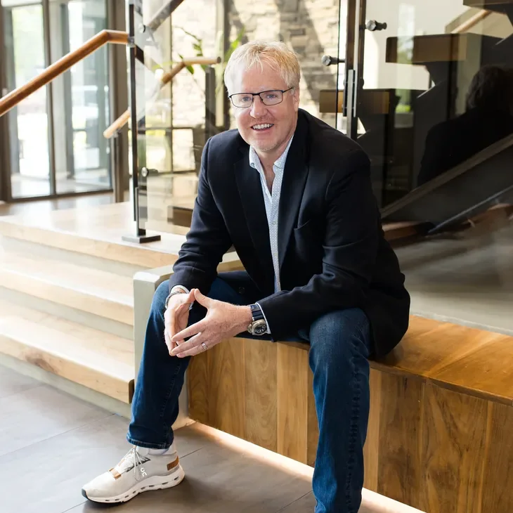 A smiling middle-aged man with glasses and blond hair, wearing a dark blazer, light blue shirt, jeans, and white sneakers, sitting on a wooden bench near stairs inside a modern building with large windows.