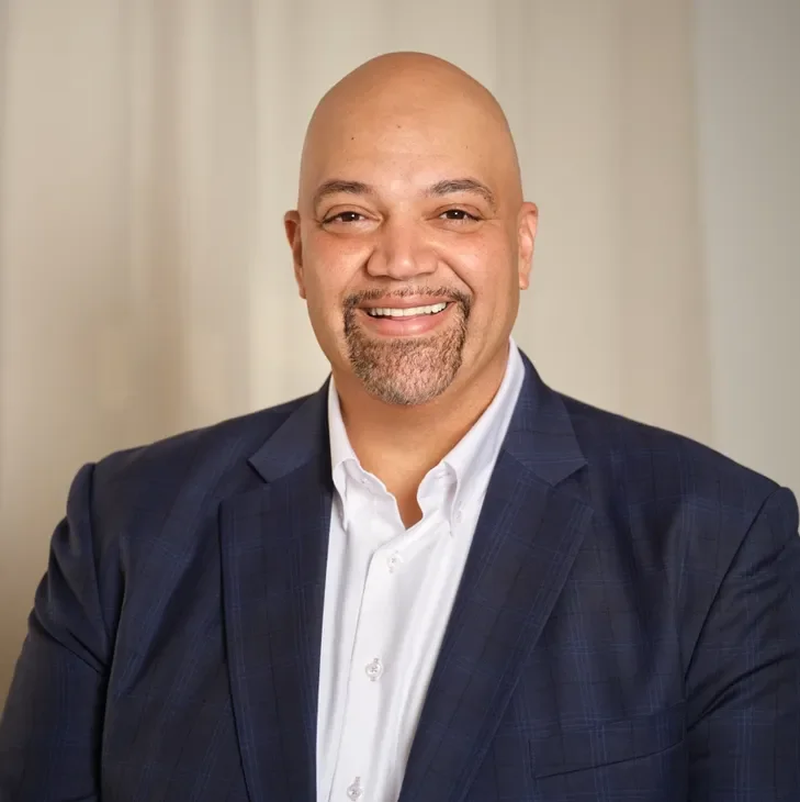 Headshot of a smiling bald man with a goatee, wearing a dark blue suit and white shirt, in a professional setting.