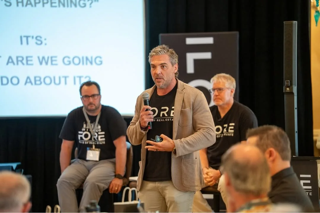 A man speaking into a microphone during a conference presentation, with two men seated behind him and a large screen displaying presentation text in the background.