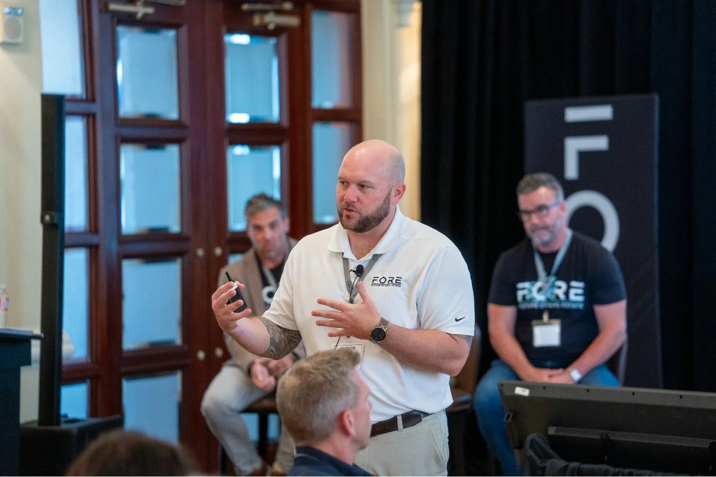 A man with a bald head, beard, and tattoos on his left arm giving a presentation at a conference. Others are seated and listening, with a large FLCC sign in the background.