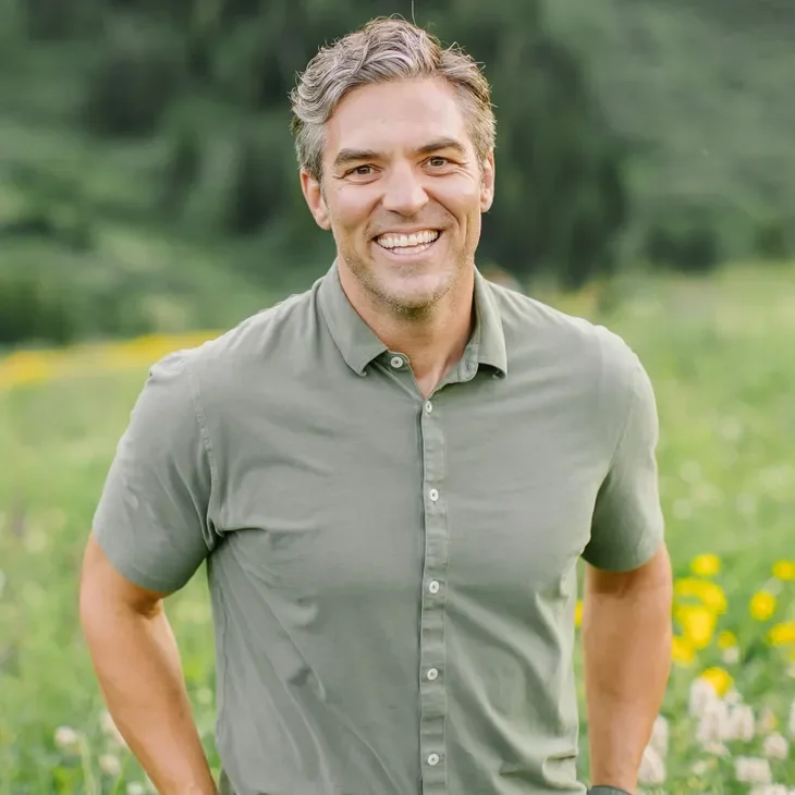 A smiling man with gray hair in a casual green button-up shirt standing outdoors in a grassy field with yellow flowers.