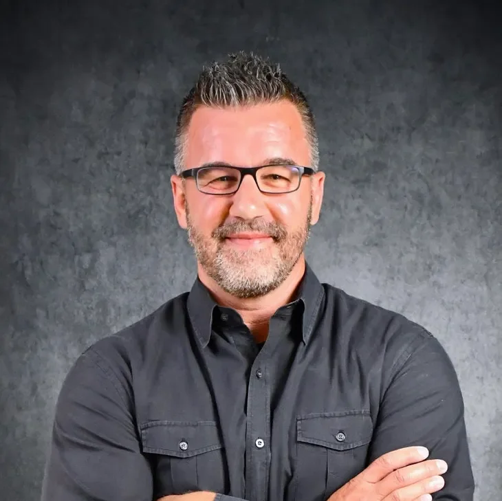 A middle-aged man with short, dark hair, beard, and glasses, smiling with arms crossed, wearing a black button-up shirt against a dark gray background.