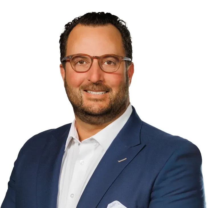 Portrait of a smiling man with glasses, wearing a navy blue suit and a white dress shirt, against a white background.
