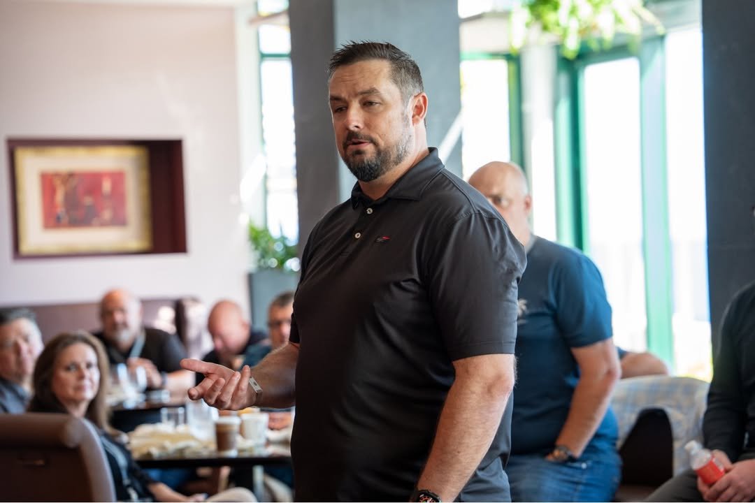 Man in black polo shirt speaking to a group in a restaurant or conference setting.