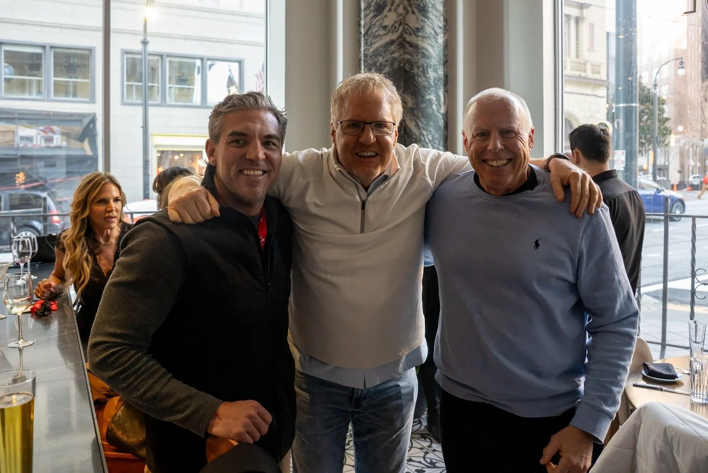 Three men smiling and posing together indoors at a restaurant or cafe, with large windows showing a busy city street outside.