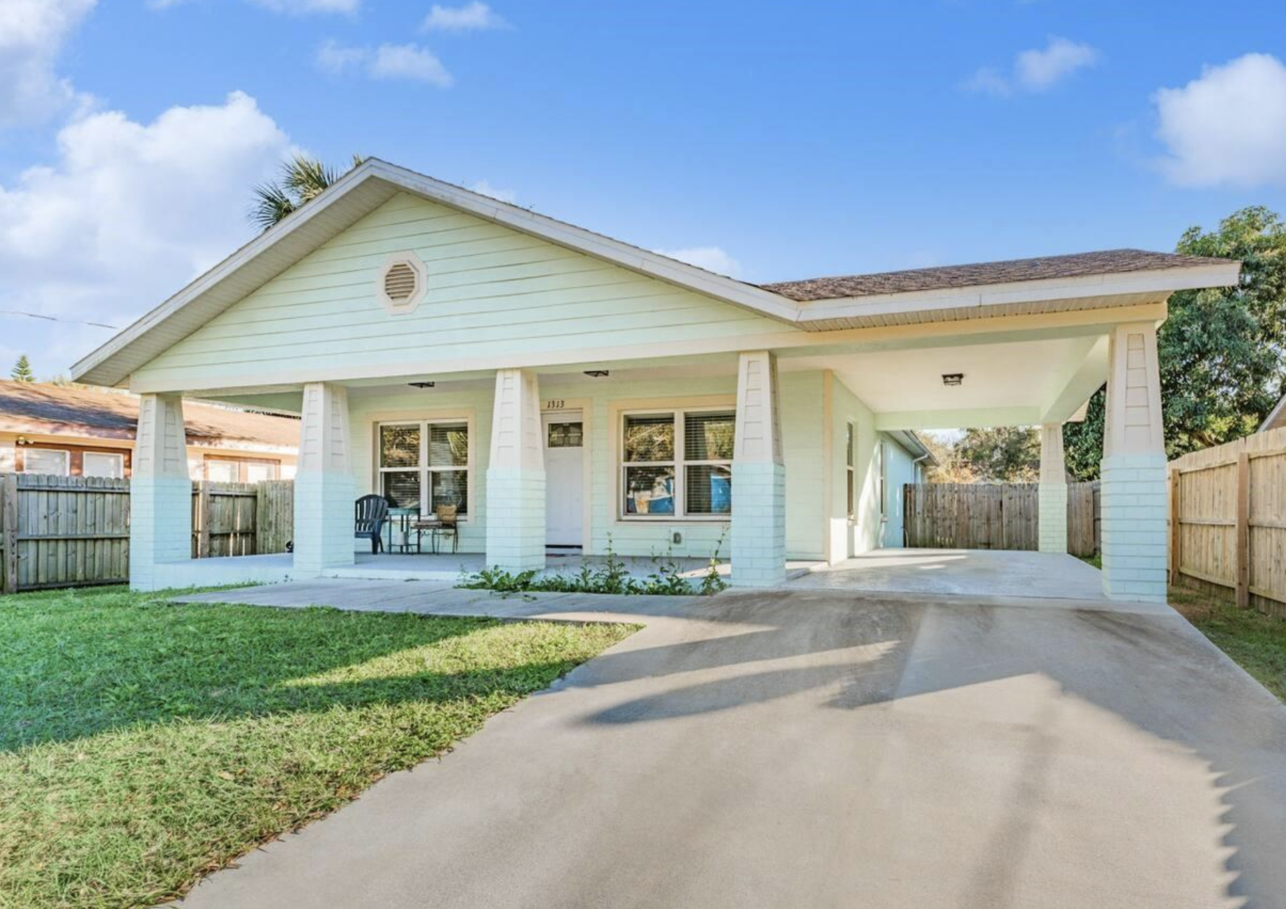 A light yellow house with a front porch, white brick columns, and a concrete driveway, surrounded by a grassy yard, wooden fence, blue sky, and a few clouds.