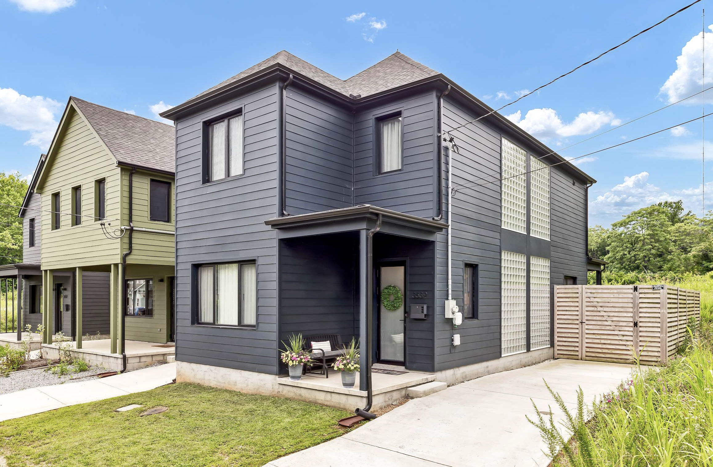 View of modern townhouse with dark blue siding, black window frames, and front porch decorated with potted plants, under a blue sky with clouds.