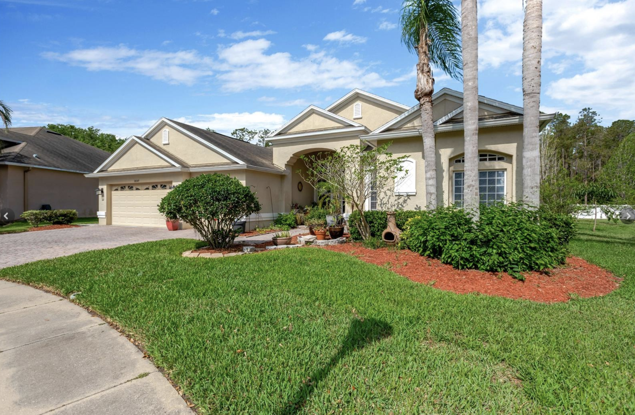 Front view of a beige house with a manicured lawn, potted plants, tall palm trees, and a bright blue sky with scattered clouds.