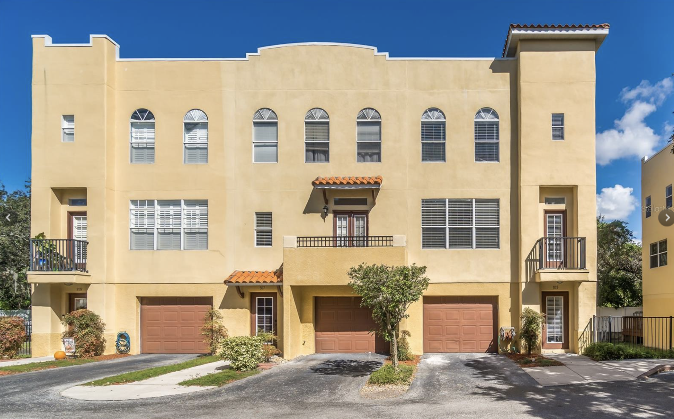 A yellow multi-story residential building with two garage doors, balconies, and multiple windows against a bright blue sky with some clouds.