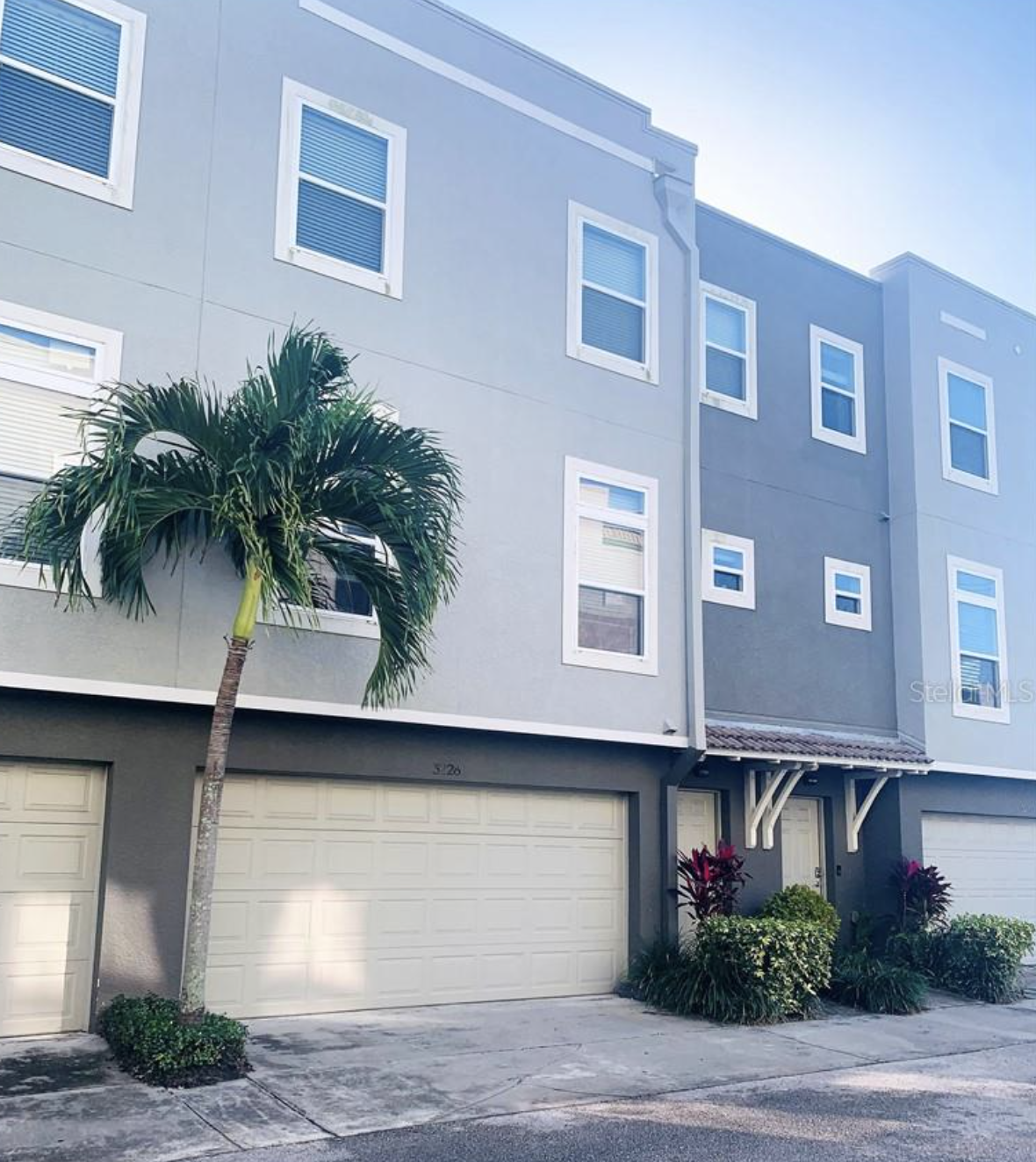 A modern, multi-story residential building painted in shades of blue and gray with several windows, a garage door, a palm tree in front, and accents of greenery near the entrance.
