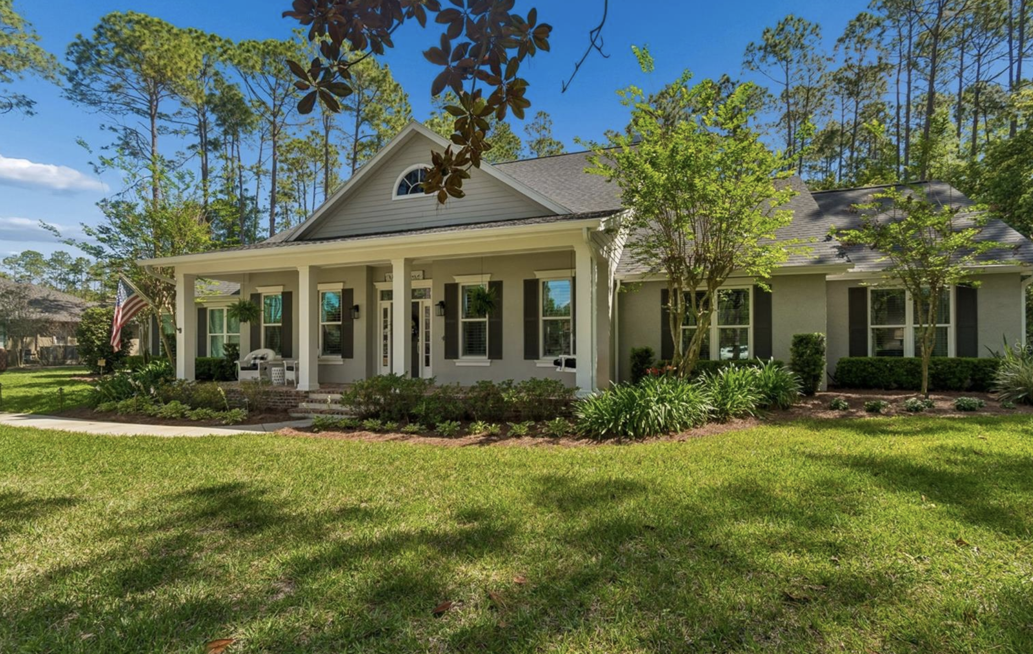 Front view of a white house with a large porch, black shutters, a gabled roof, surrounded by green trees and a well-maintained lawn, under a blue sky.