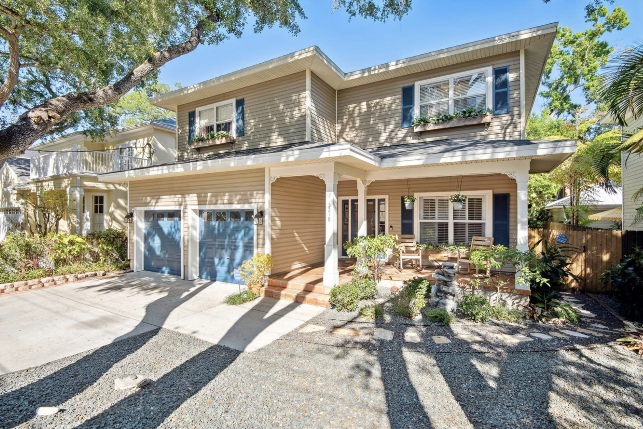 Front view of a beige two-story house with blue shutters, a small porch, and a two-car garage. There are plants and small trees in the front yard and a large tree shadow over the driveway on a sunny day.