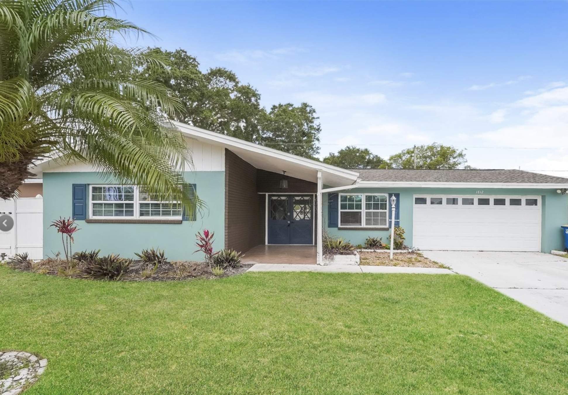 A single-story house painted in light blue with dark blue shutters and a white garage door. The front yard has green grass, small red and green plants, and a palm tree on the left side. The house has a small covered porch with a blue front door and a window to the left of it, and another window to the right. The house number '1812' is visible above the garage.