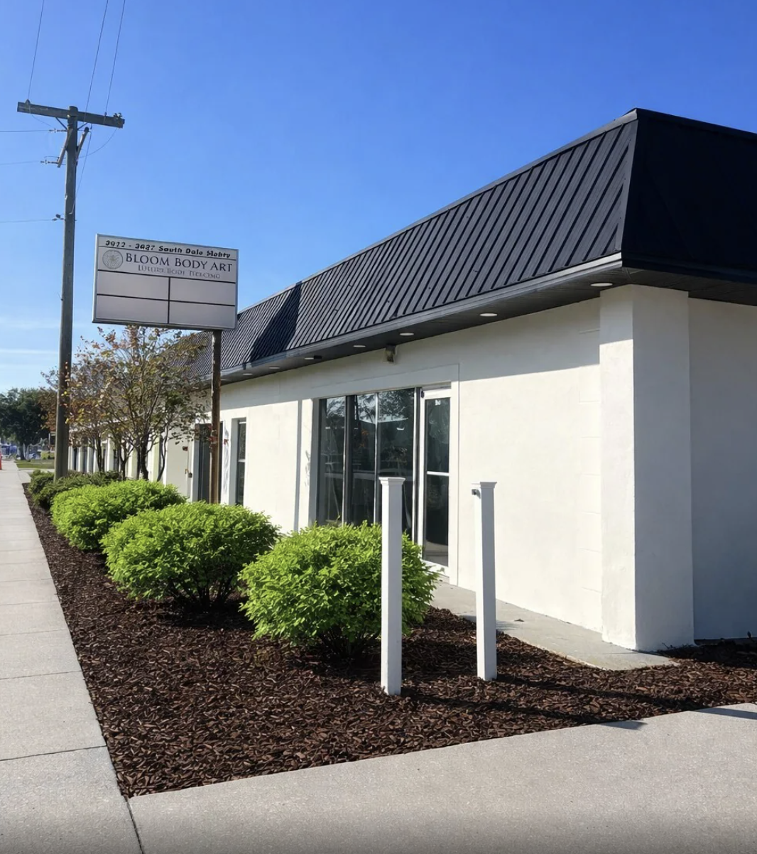 A white building with large windows and a black metal roof, featuring a sidewalk and well-maintained green bushes in front. There is a signboard with the text 'Bloom Body Art,' and a utility pole with electrical wires nearby. The sky is clear and blue.