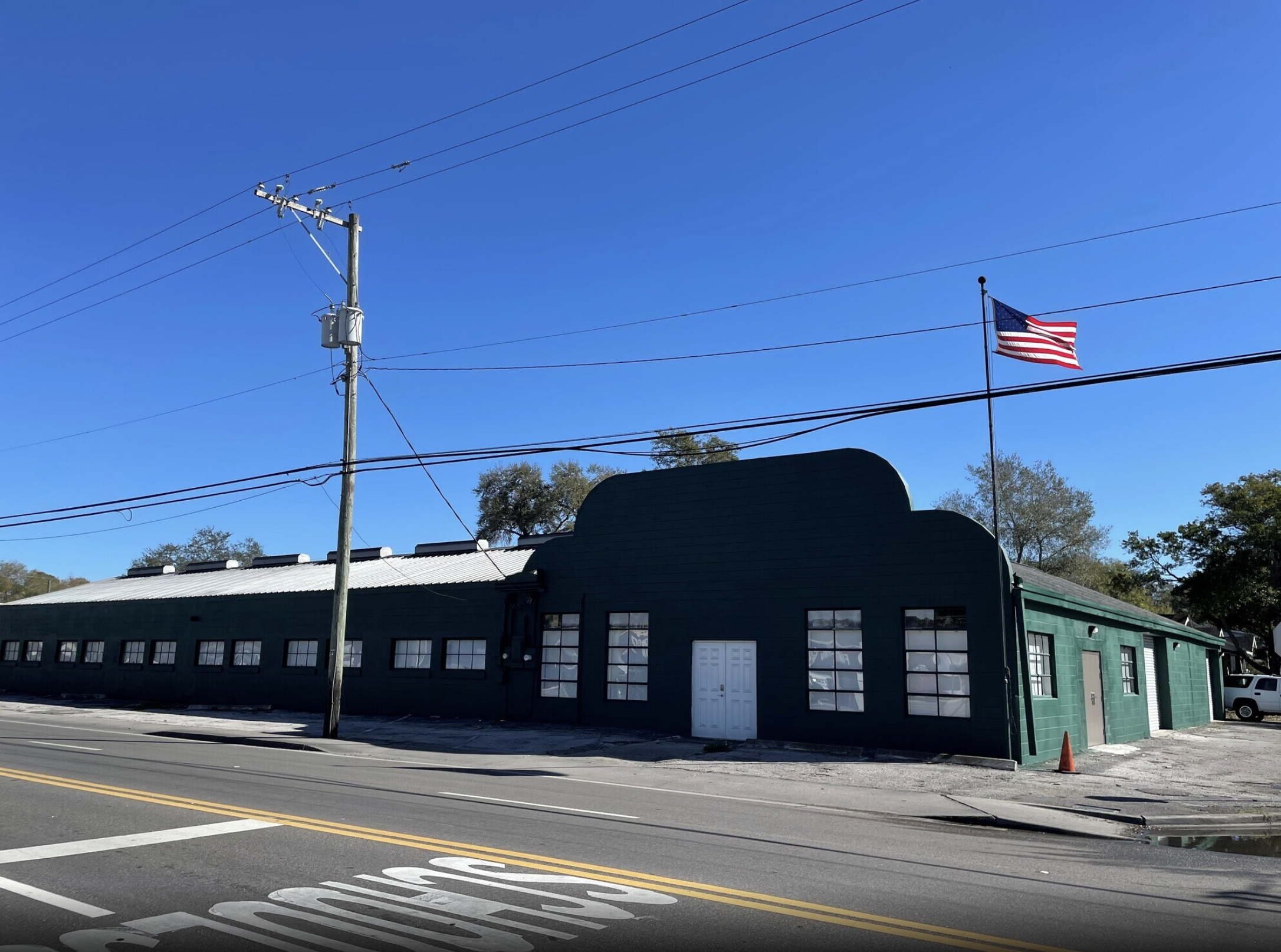 A street scene with an industrial-style building painted dark green, a utility pole with power lines, an American flag on a flagpole, and a clear blue sky.