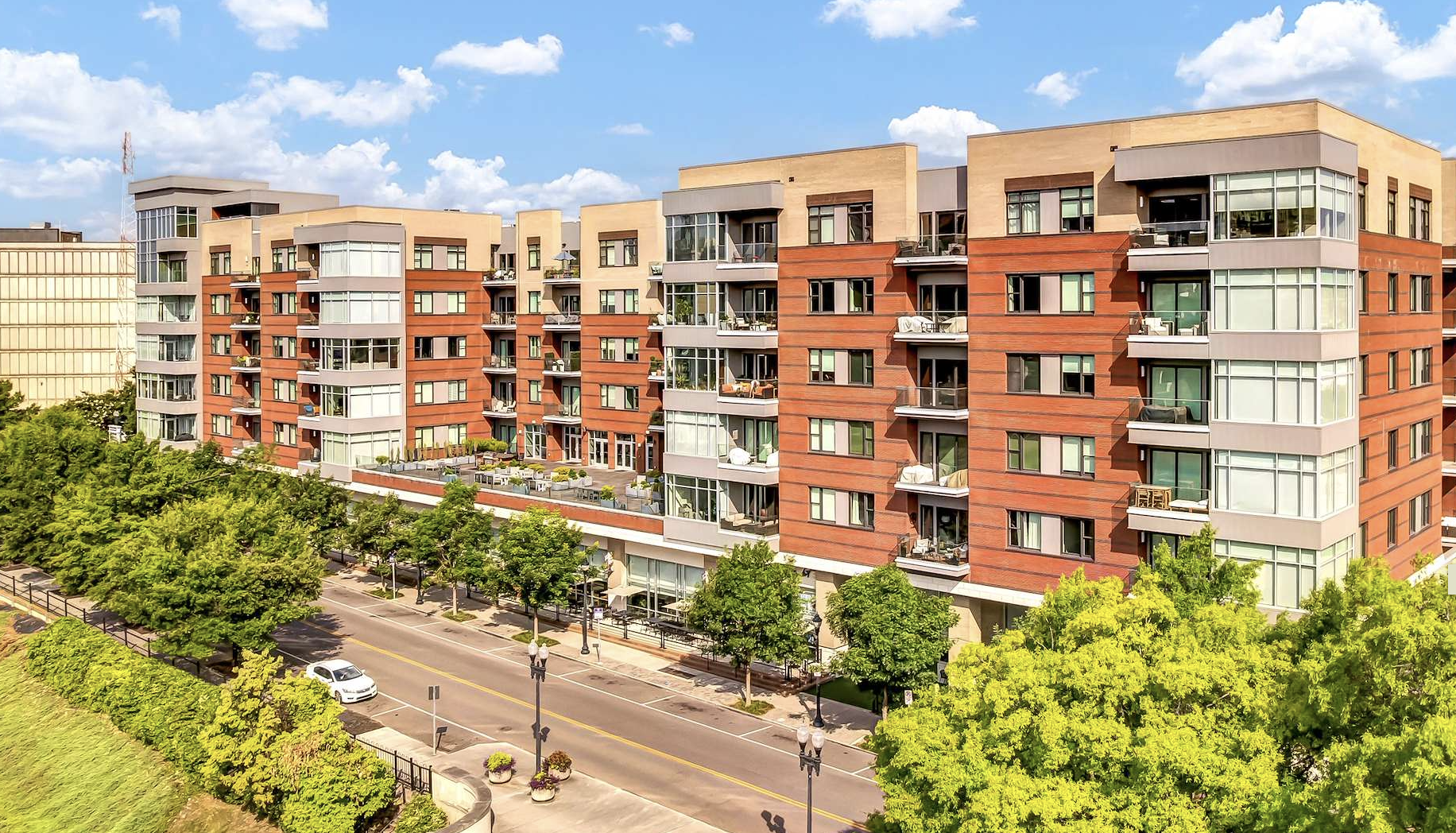 A modern multi-story apartment building with large windows and balconies, surrounded by trees and a street with parked cars and street lamps.