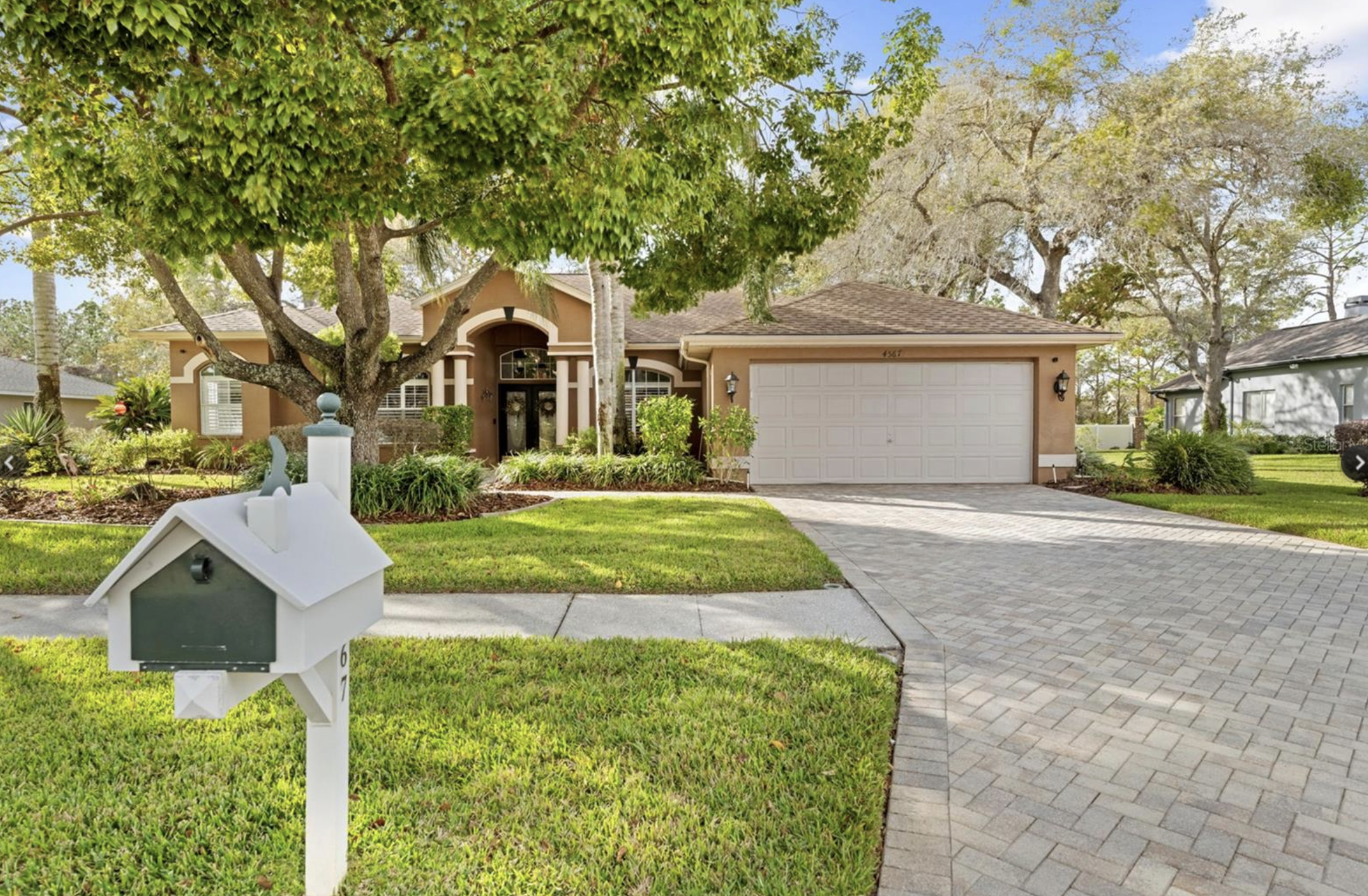 Front yard of a house with a large tree, a white mailbox, a driveway, and a tan house with a garage and a black front door surrounded by greenery.