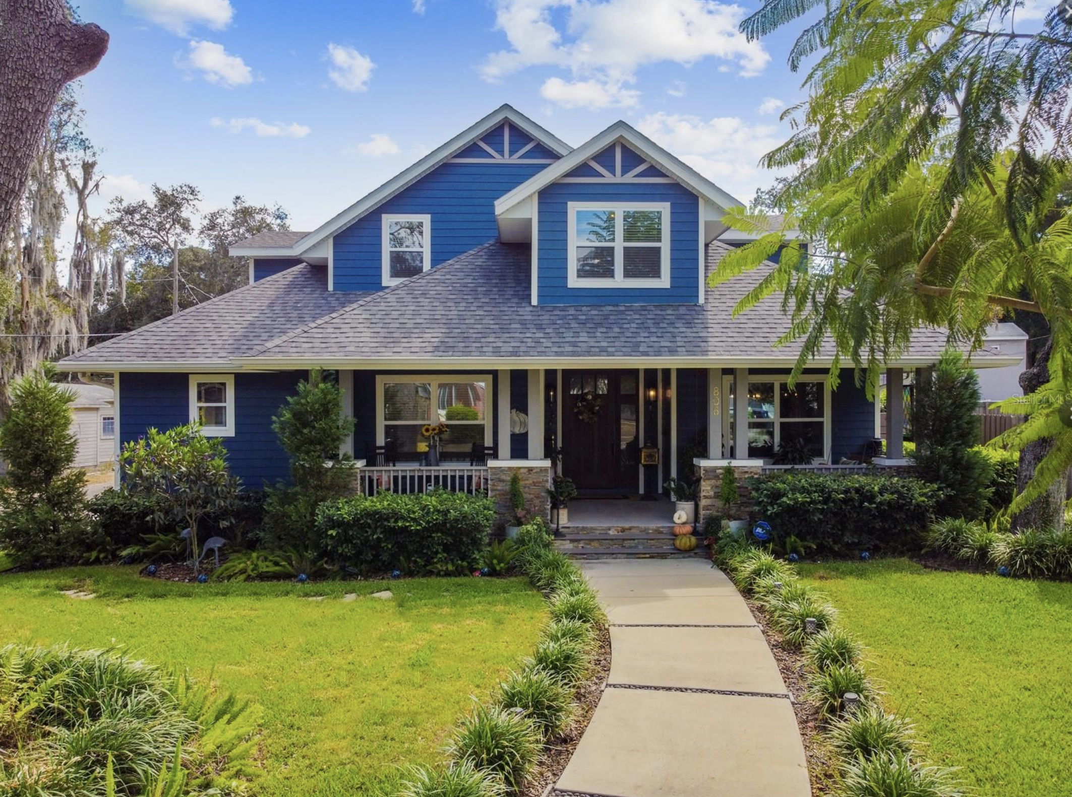 A blue, two-story house with grey roof and white trim, surrounded by lush green plants and trees, with a front walkway leading to the porch.