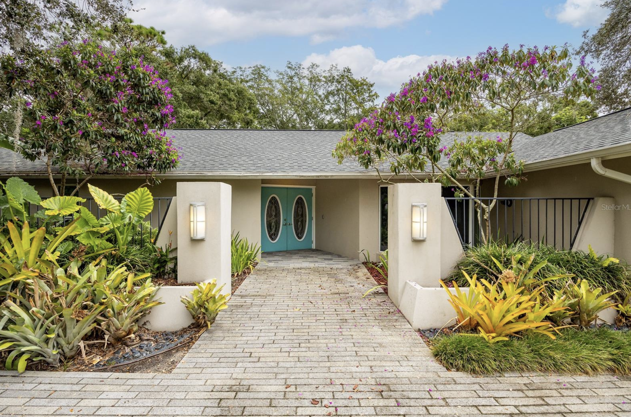 A house with a grey shingle roof, teal double doors, and white walls. A paved walkway leads to the entrance, flanked by lush green and yellow plants. Two flowering trees with purple blossoms are on either side of the house. The sky is partly cloudy with patches of blue.