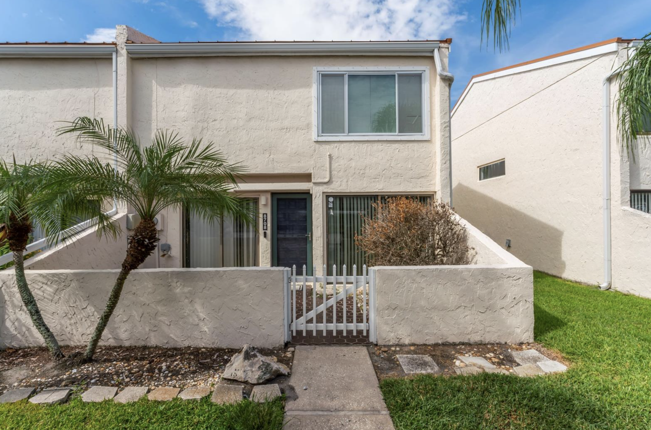 Front view of a two-story beige house with a white fence, palm trees, small bushes, and a brick walkway at the entrance, under a partly cloudy sky.