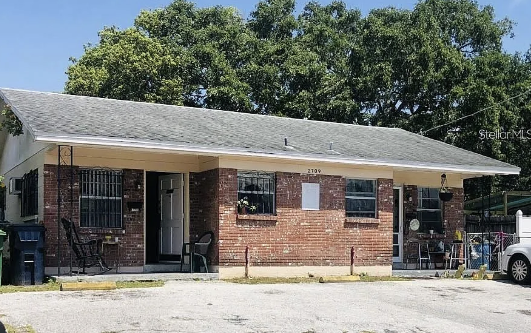 A small brick house with a gray sloped roof and a porch. The house has two large windows with black security bars and a white front door. There are items like chairs, hanging plants, and a small table on the porch. The house number 2709 is visible above the porch. Behind the house, large green trees and a clear blue sky can be seen.