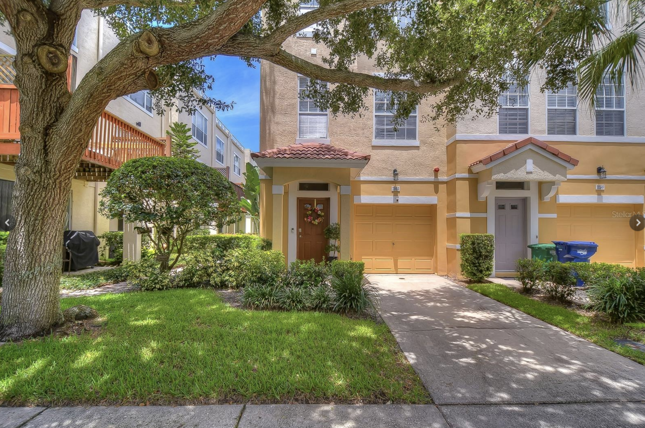 Front yard of a multi-story yellow townhouse with brown garage door, surrounded by green bushes and trees, with a sidewalk in the foreground.
