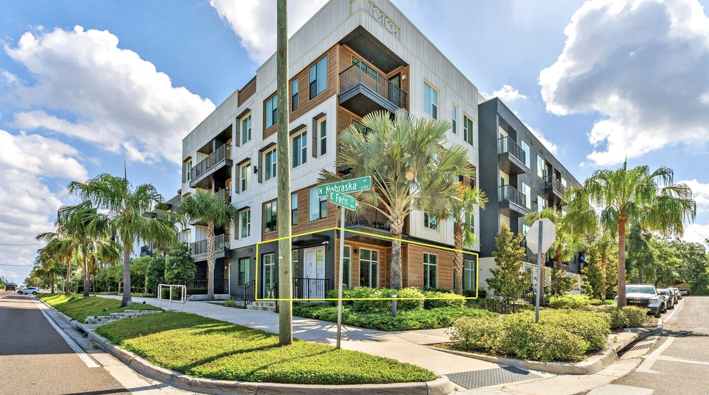 Modern multi-story apartment building on corner of N Nebraska Avenue and E Fern Street, with palm trees and a sidewalk in the foreground, under a partly cloudy sky.