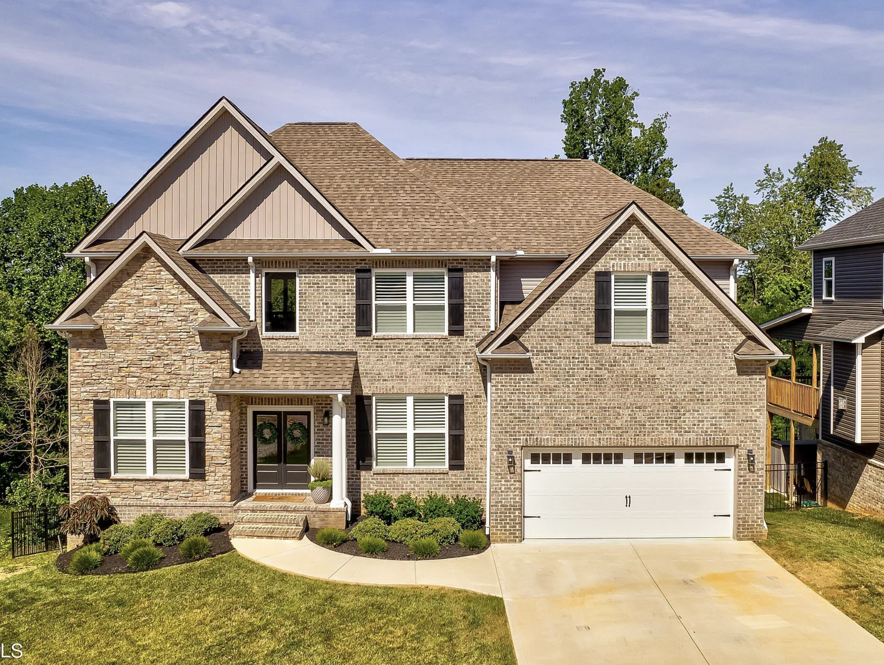 A two-story house with brick and siding exterior, large windows with black shutters, a front porch with steps, and a white garage door. The front yard has a curved walkway, green grass, shrubs, and landscaped plants. There are trees and neighboring houses visible in the background, under a partly cloudy sky.