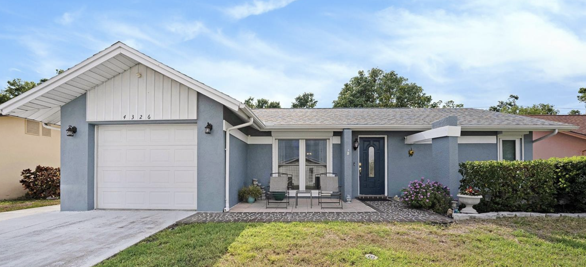 A single-story house with a blue exterior, white garage door, front porch with two chairs, and a landscaped yard with bushes and flowers. Blue sky with some clouds in the background.
