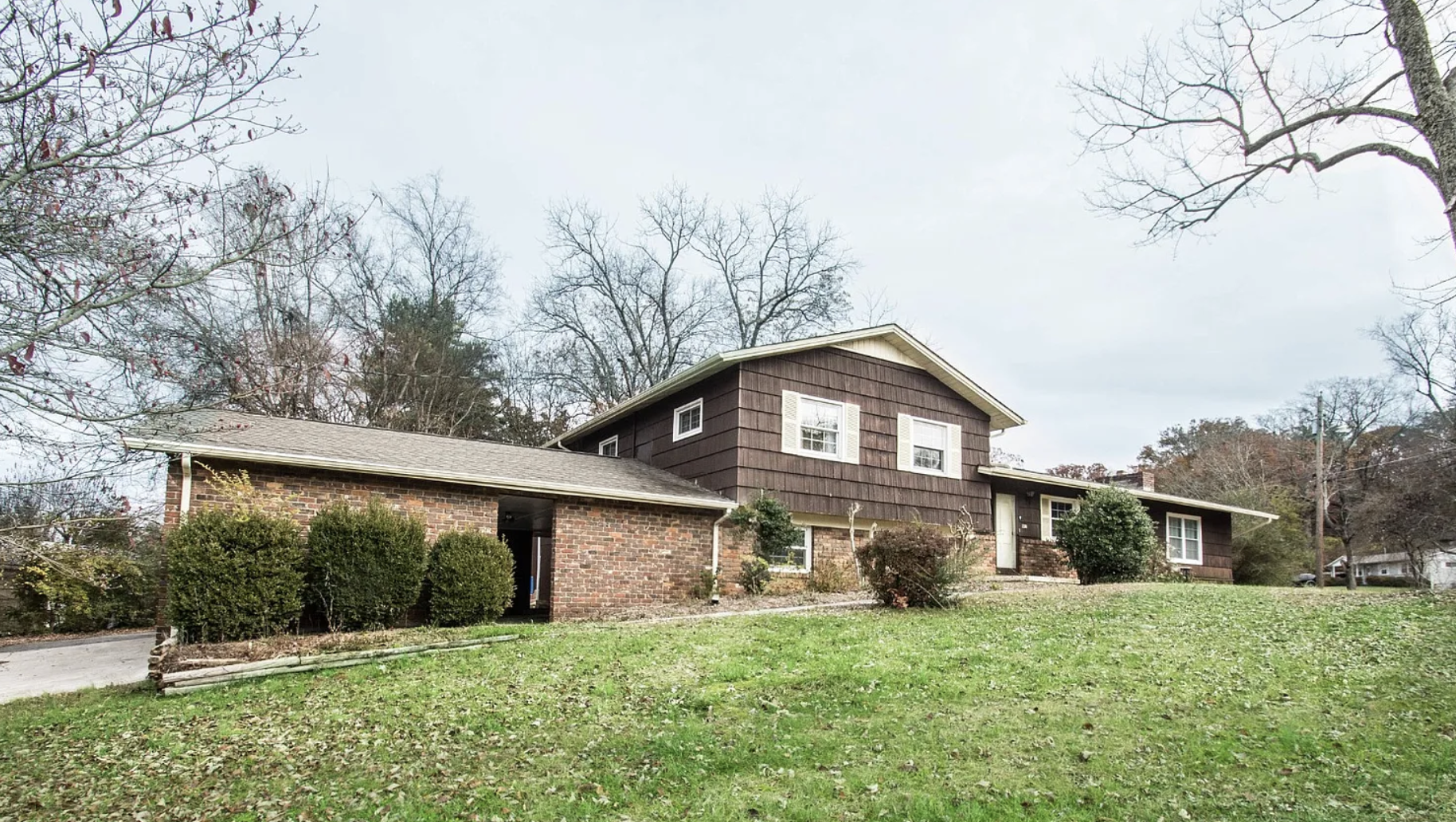 A two-story house with a brick and brown wooden exterior, surrounded by a grassy yard and leafless trees under an overcast sky.