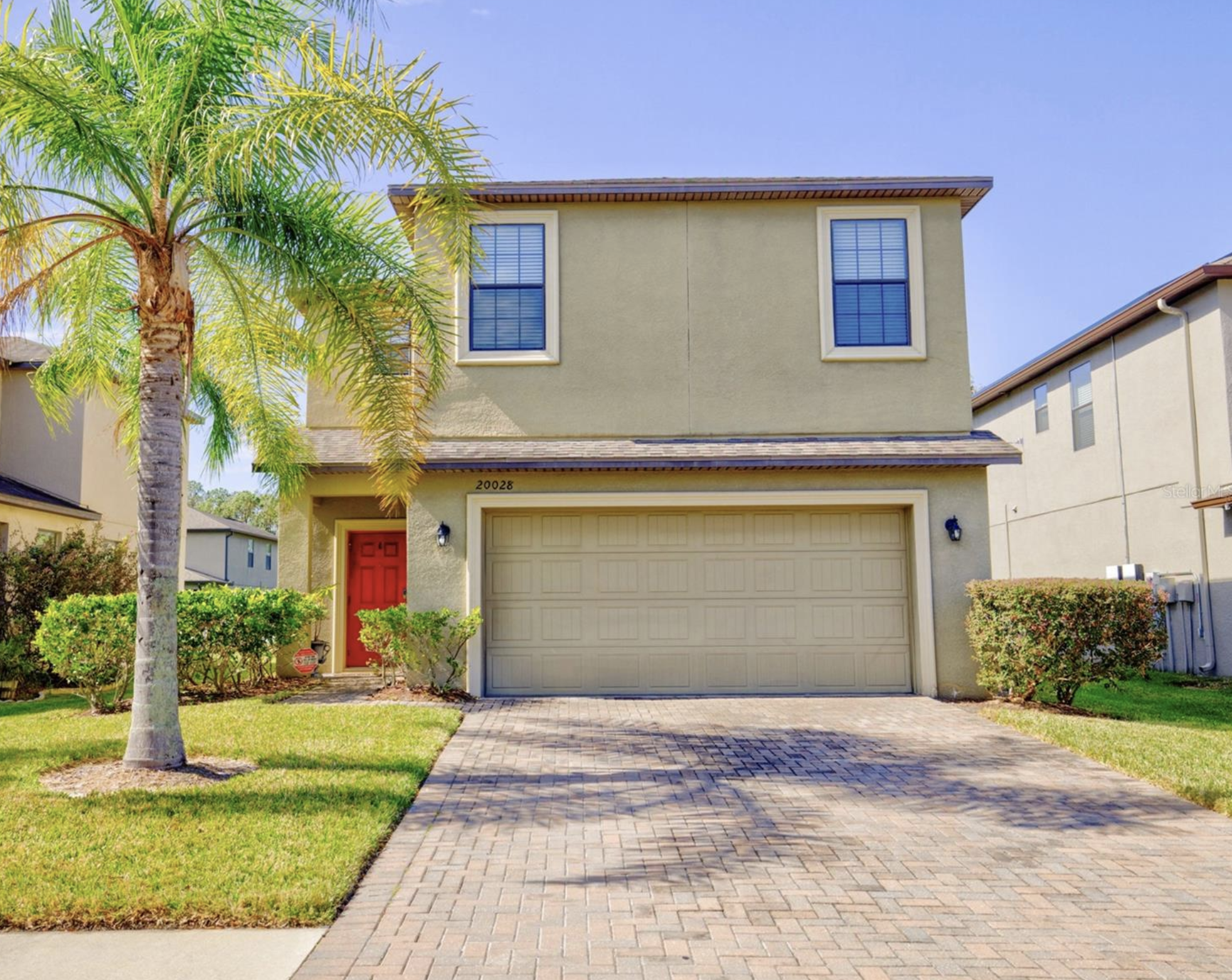 A two-story house with beige walls and a red front door. It has double windows on the upper level and a garage door on the lower level. There is a driveway made of paver bricks leading up to the garage. A palm tree and green bushes are in front of the house, with a blue sky in the background.