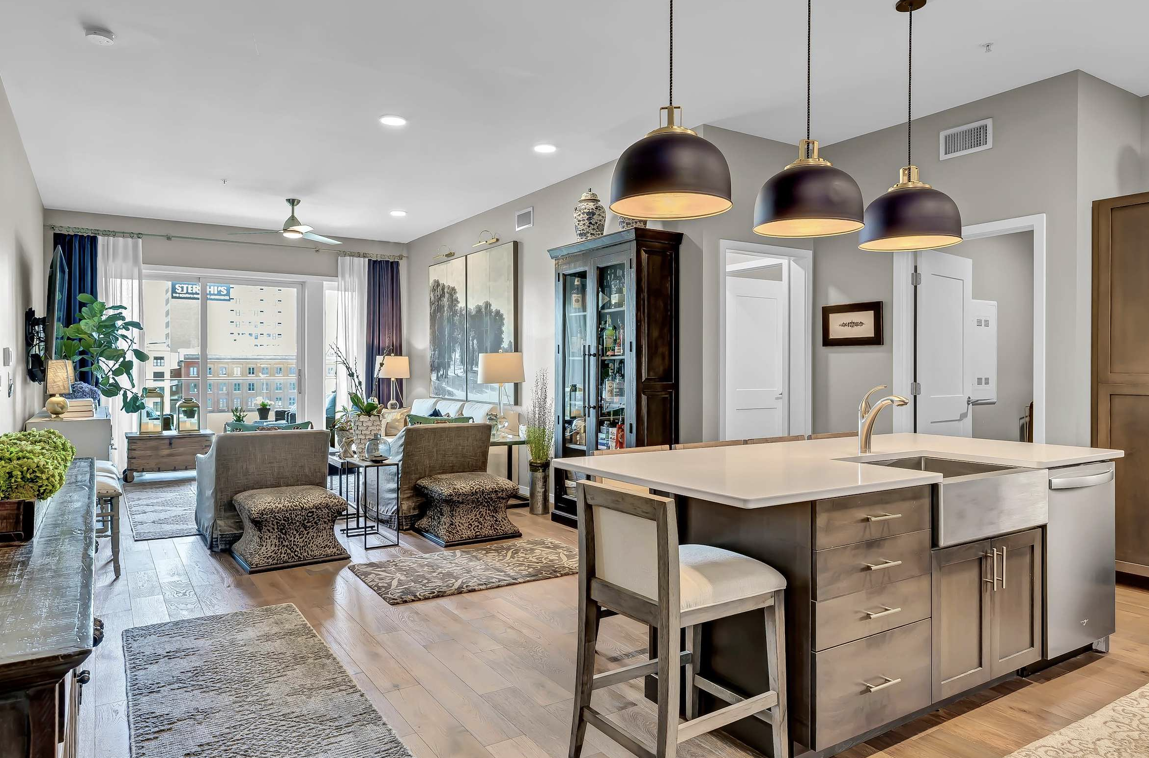 Living room and kitchen area in an apartment with modern decor. There are three black pendant lights hanging above the kitchen island, a large painting on the wall, a glass-front cabinet, and a sliding glass door leading to a balcony with city views.