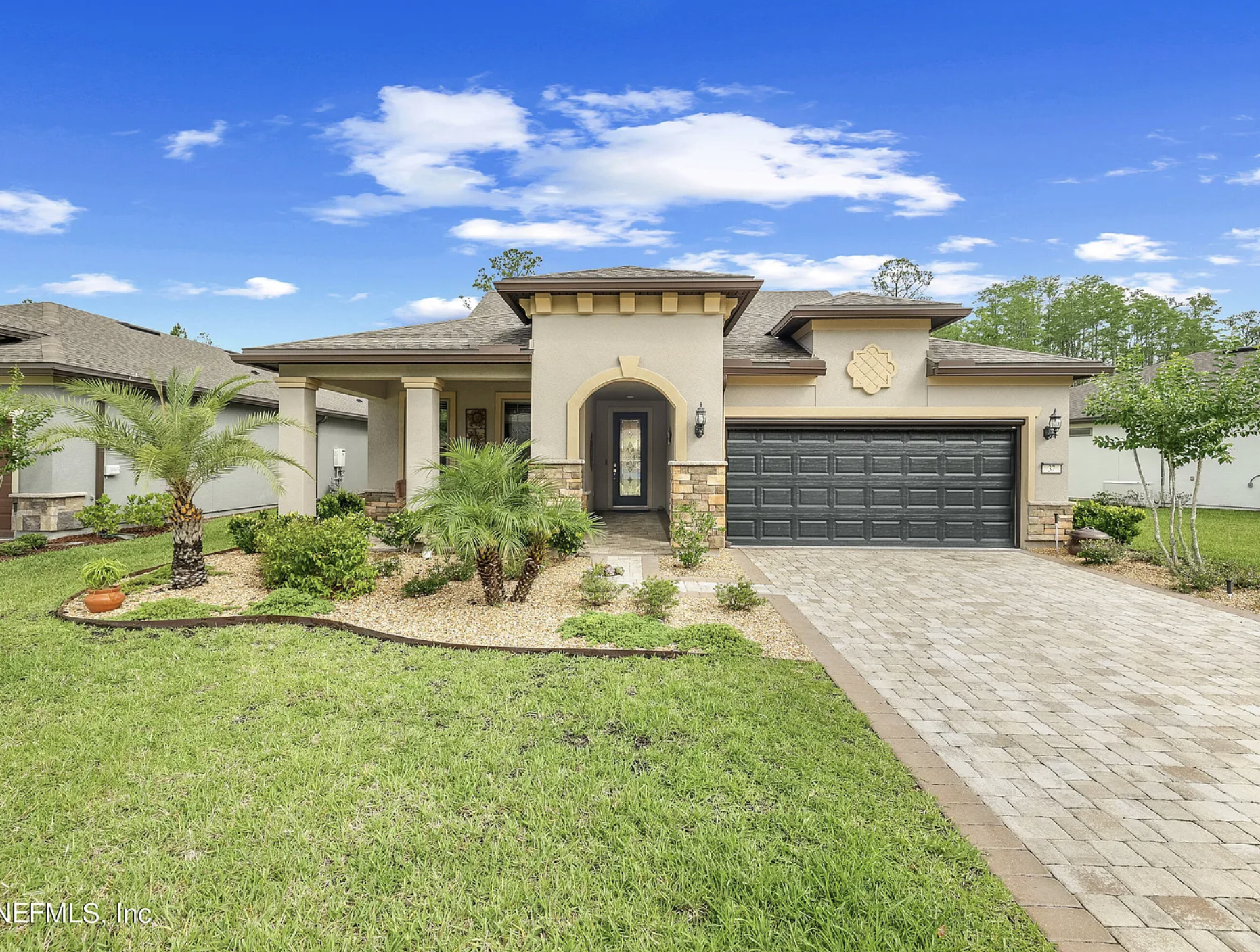 A modern single-story house with a well-maintained front yard featuring palm trees and lush green grass. The house has a gray garage door, a decorative arched doorway, and a stone pathway leading to the entrance. The roof is shingled, and the house has decorative trim and wall accents. The sky above is blue with a few scattered white clouds.