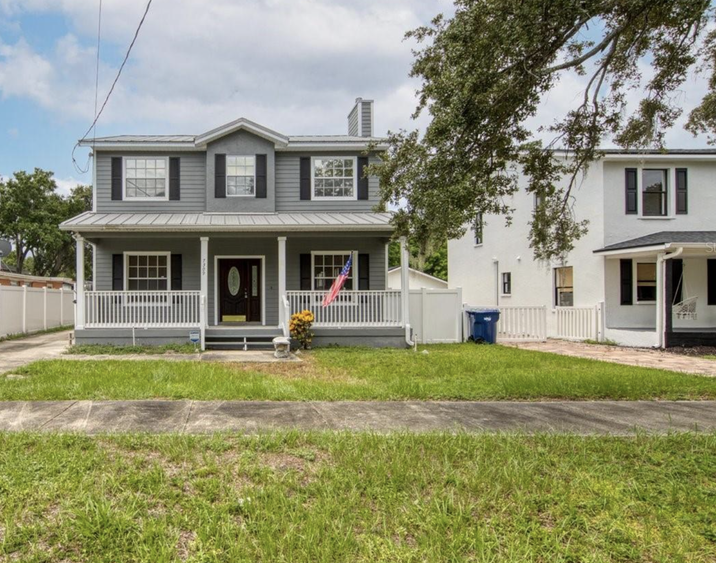 Two-story gray house with black shutters, front porch with white railing, American flag, and a flower pot with yellow flowers in the front yard.