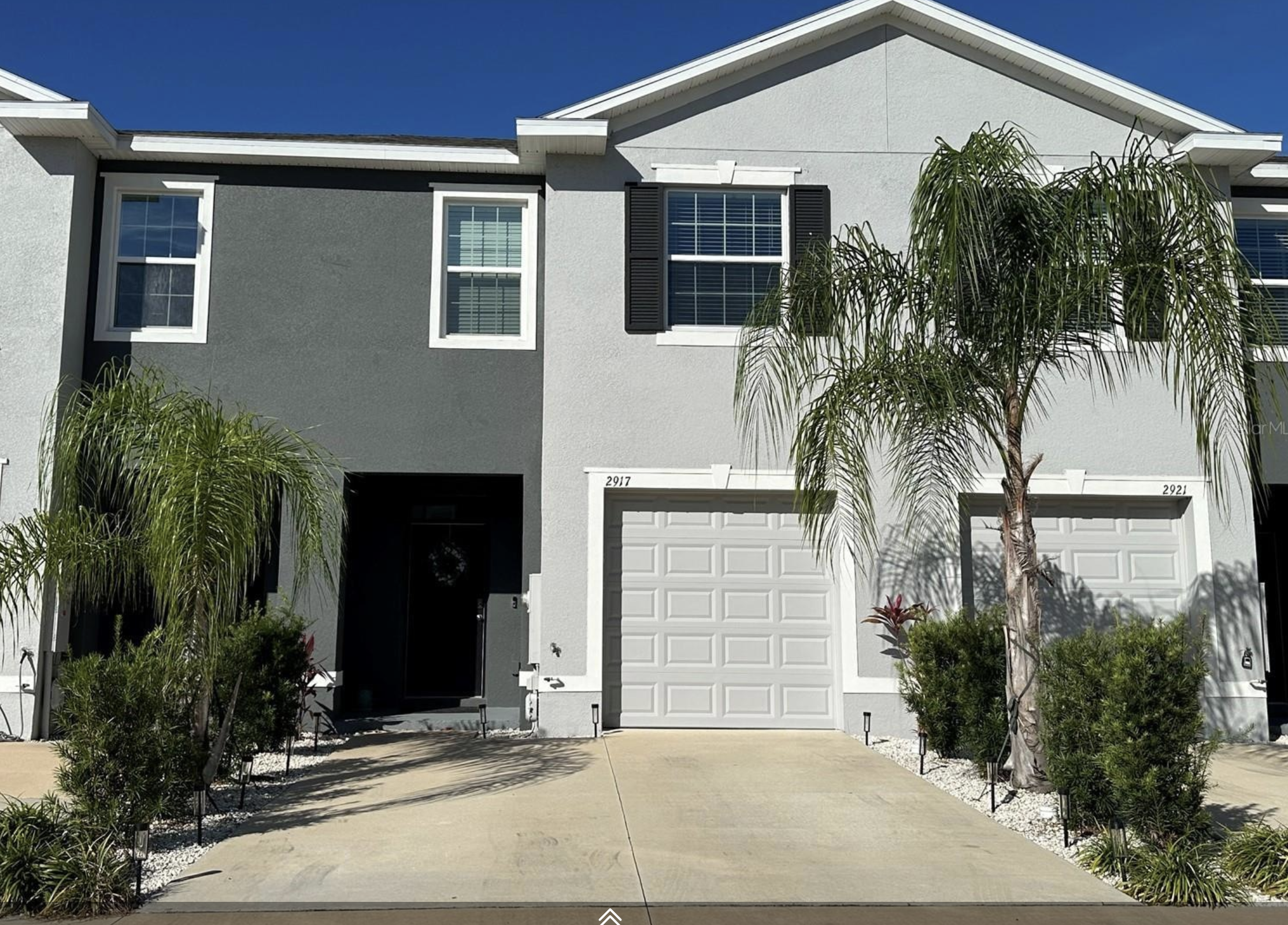 Front view of modern two-story house with white garage door, black window shutters, and trees in front yard under clear blue sky.