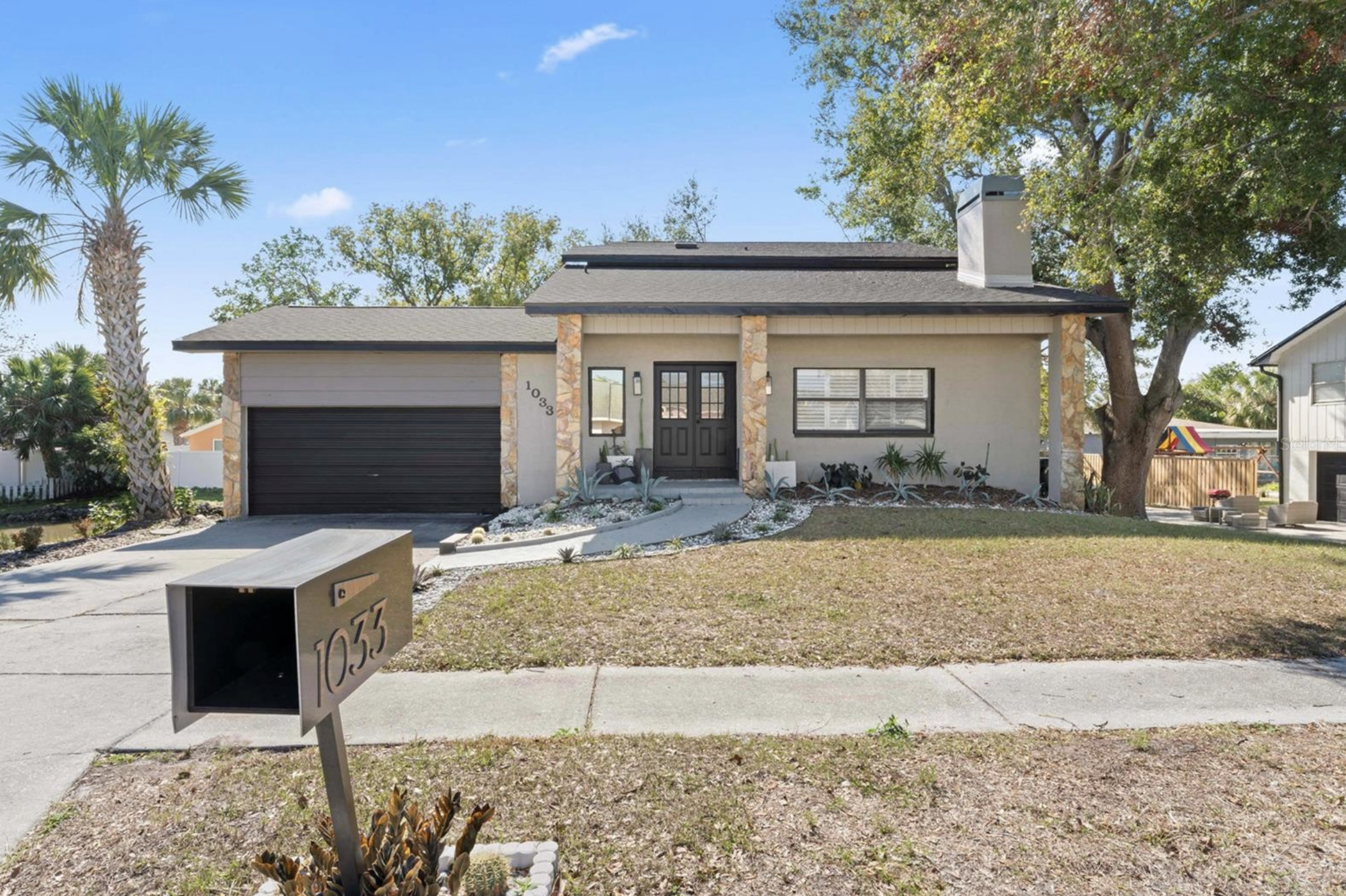 A modern single-family house with a gray exterior, stone accents, and a black garage door. There is a front porch with a door, windows, and landscaped yard with succulents and plants. A mailbox with house number 1033 is in the foreground.