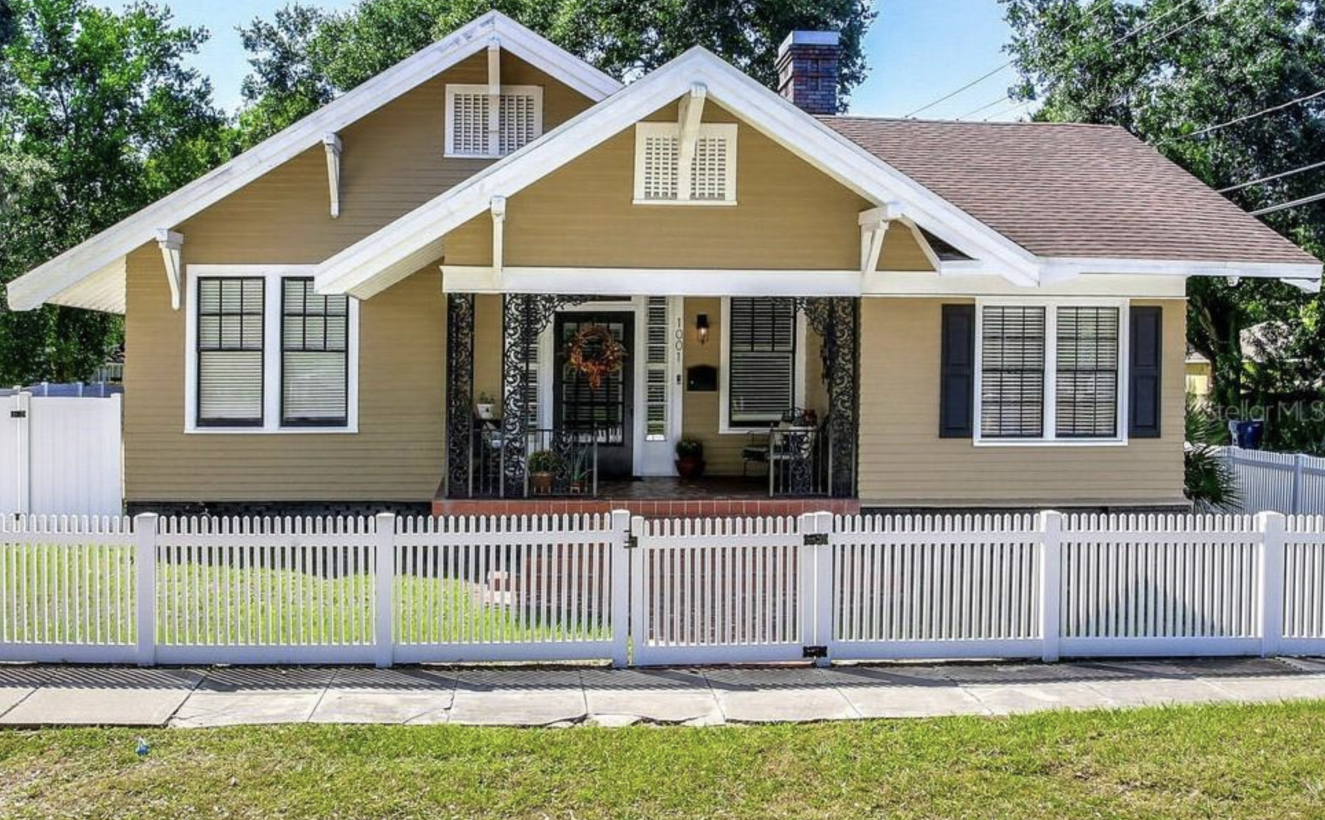 A yellow house with a white picket fence, metal decorative posts on the porch, and large windows, surrounded by trees under a clear sky.