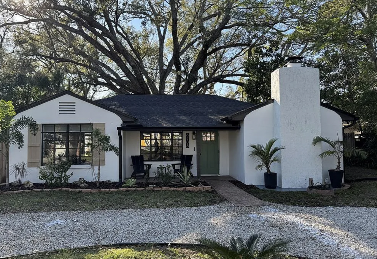 A small white house with black roof shingles, a green front door, and a chimney. The house has a front porch with two black chairs and plants in pots and in the ground. Tall trees and a gravel driveway are in front.