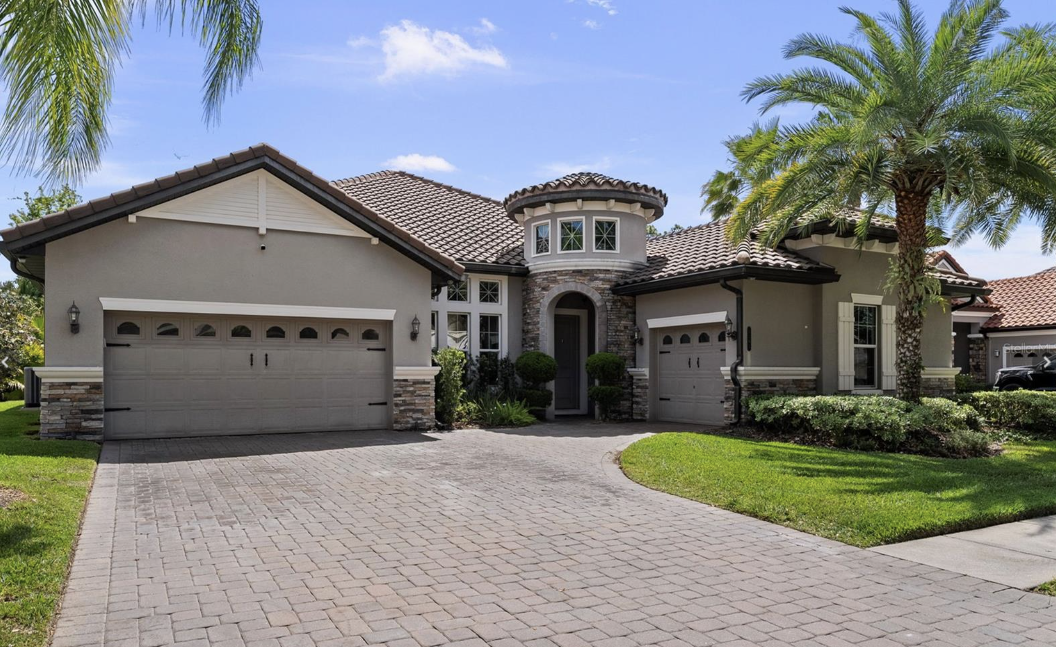 A modern two-story house with a gray front door, two garage doors, and a tiled roof, surrounded by a green lawn and palm trees under a blue sky.
