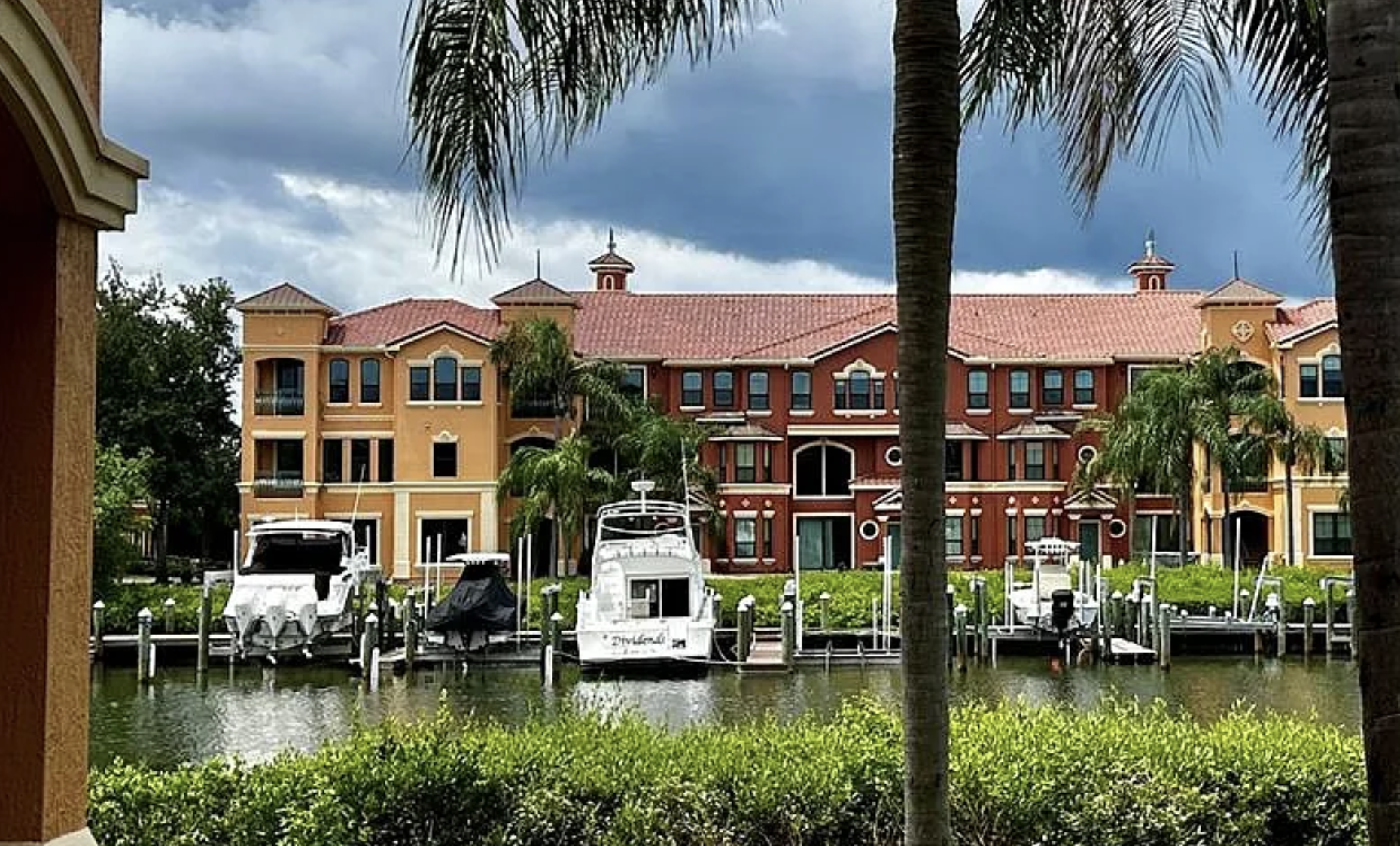 View of colorful multi-story apartment buildings with boats docked along a marina, framed by palm trees and lush greenery, with cloudy sky overhead.