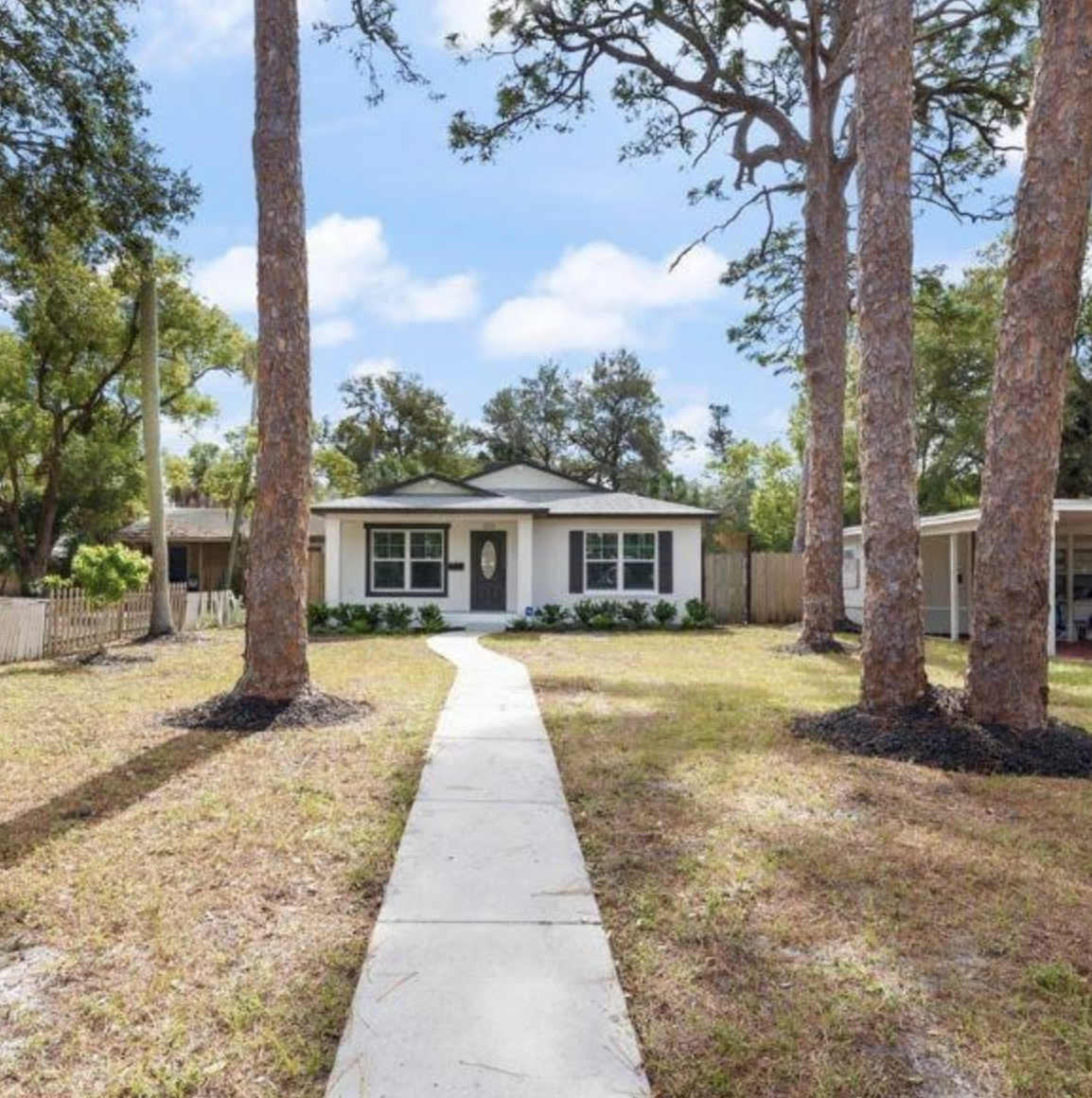 A white single-story house with black shutters, a dark door, and a curved walkway leading to the front porch, surrounded by tall pine trees in a yard with sparse grass and a partly cloudy sky.