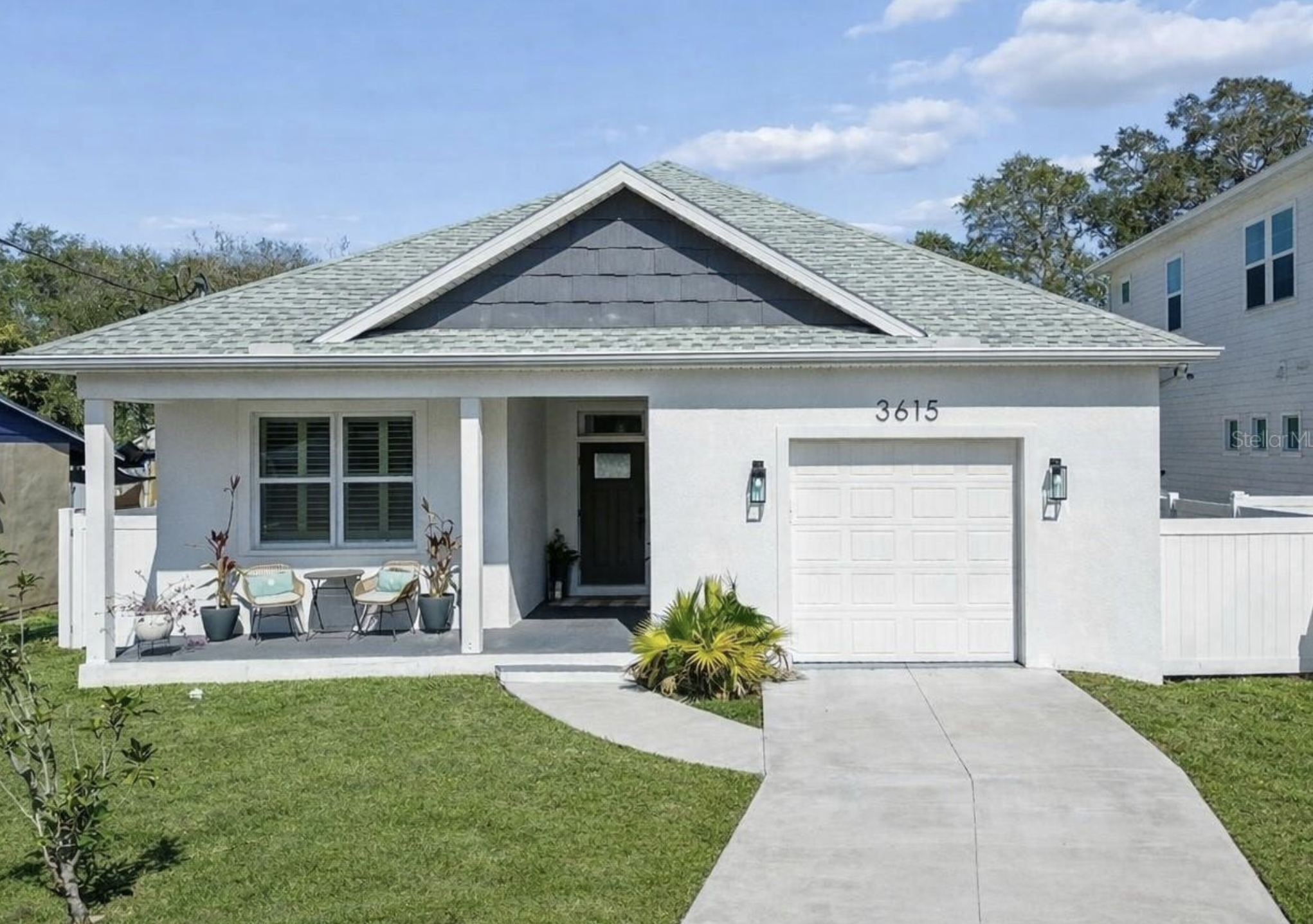 White house with grey roof, front porch with patio chairs, green lawn, and sidewalk, clear sky with some clouds, house number 3615.